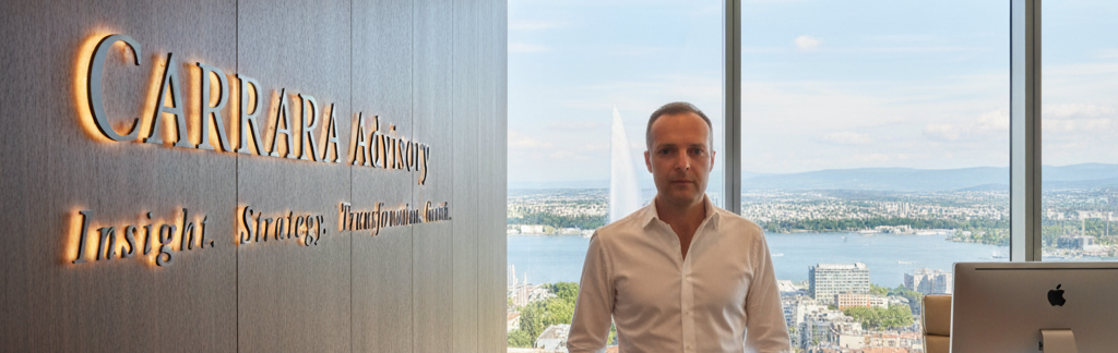A man standing in an office with a view of a city and river through large windows, with a sign on the wall that reads 'CARRARA Advisory' and a tagline 'Insight. Strategy. Transformation. Change.'