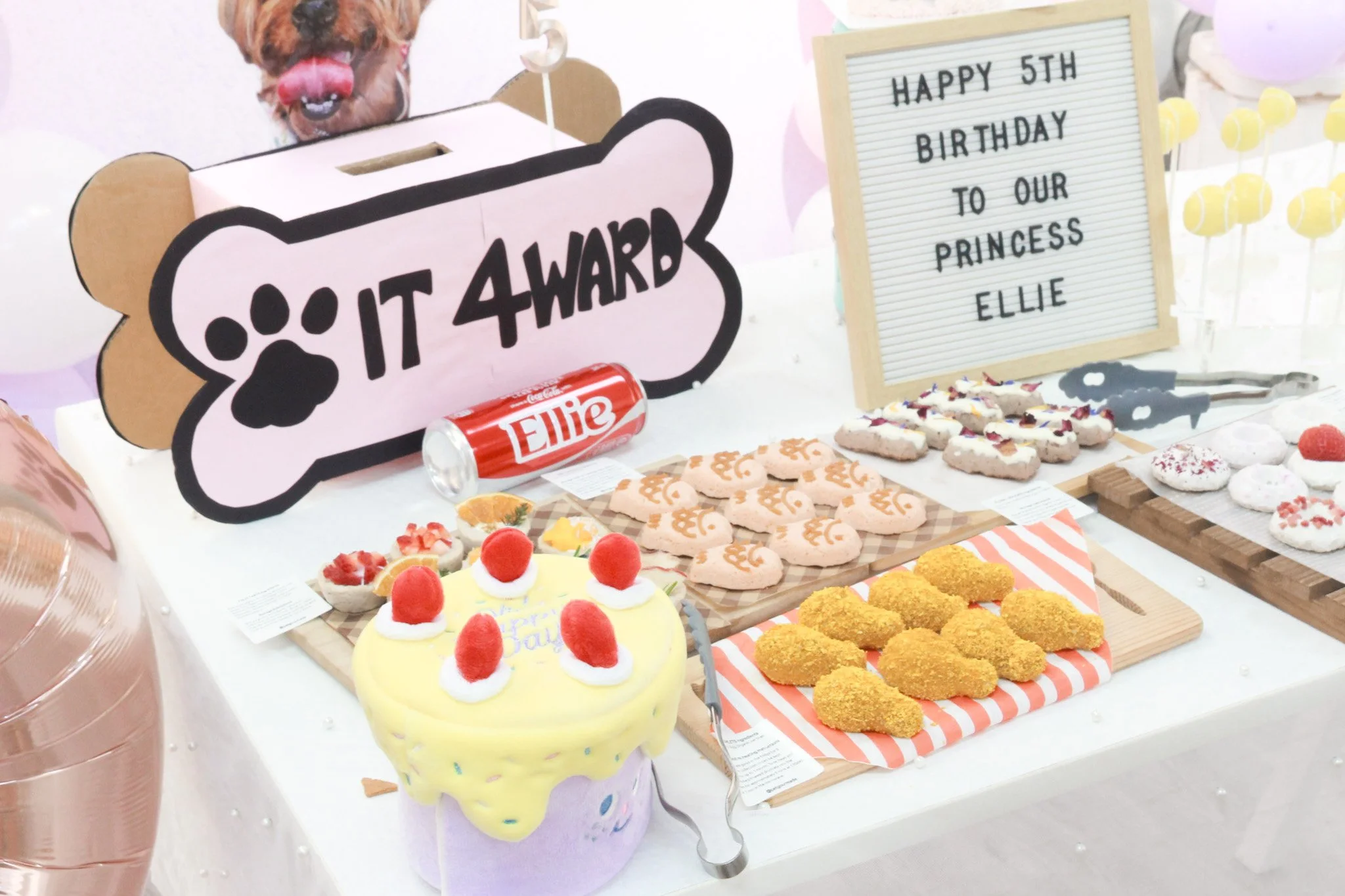 Cake, cookies, and fried snacks on a table for a dog-themed birthday celebration. A sign reads "IT 4 WARD" with a pet paw print, and a letter board says "HAPPY 5TH BIRTHDAY TO OUR PRINCESS ELLIE."