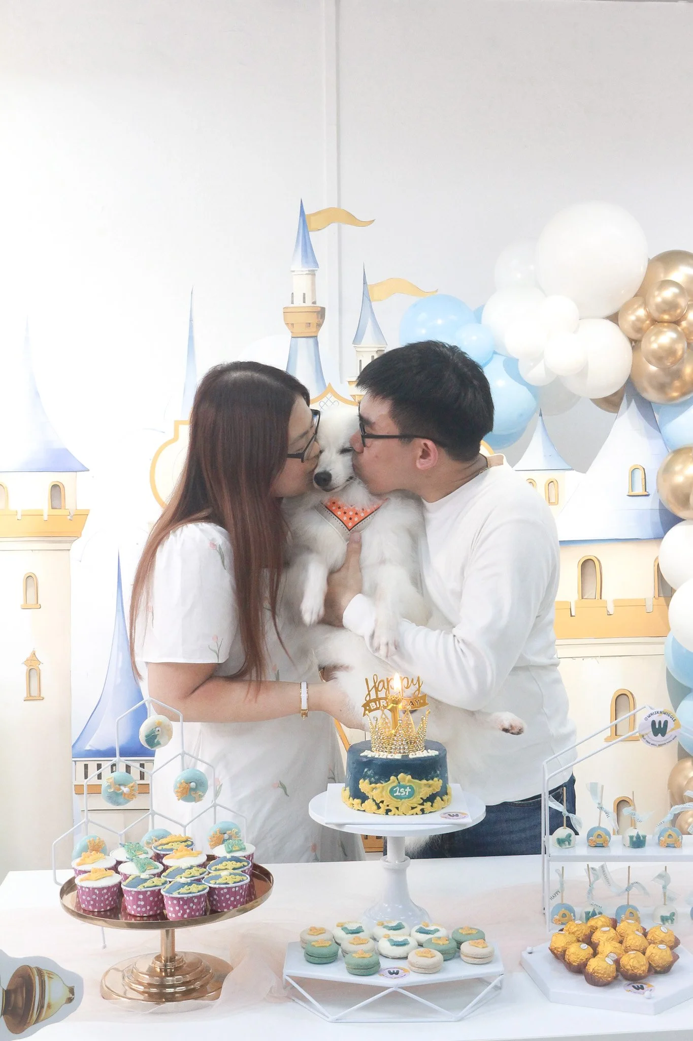 A couple dressed in white are kissing a dog with a birthday cake and treats on a table, in front of a fairy tale castle backdrop with balloons.