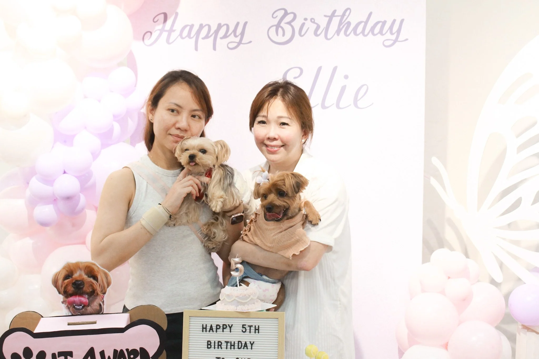 Two women celebrating a dog's fifth birthday with puppies, a cake, and pastel balloons at a themed birthday party.