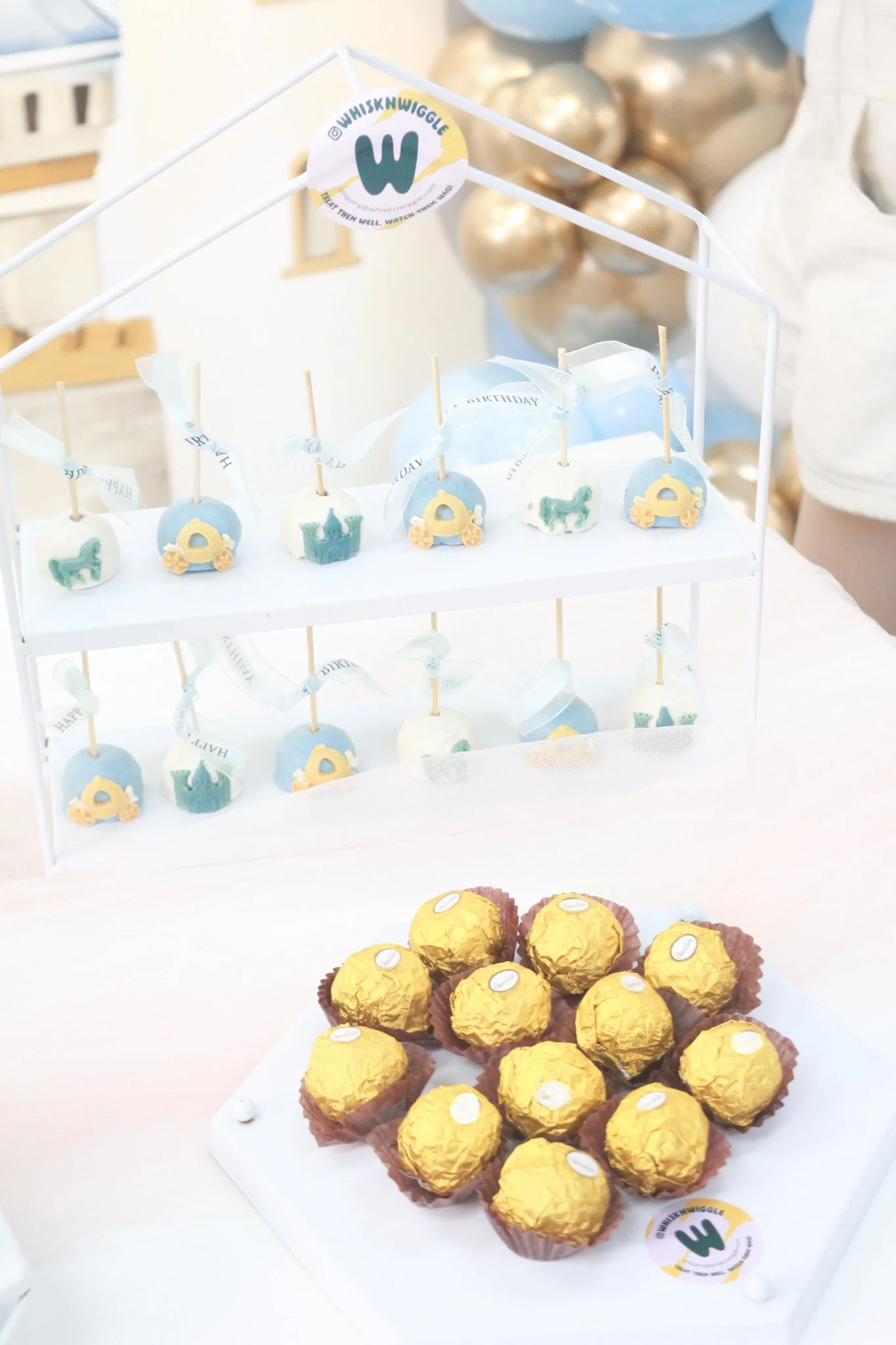 A display of decorated cake pops with blue and white icing on a white tiered stand, and chocolate truffles covered in gold foil on a white plate at a birthday celebration.