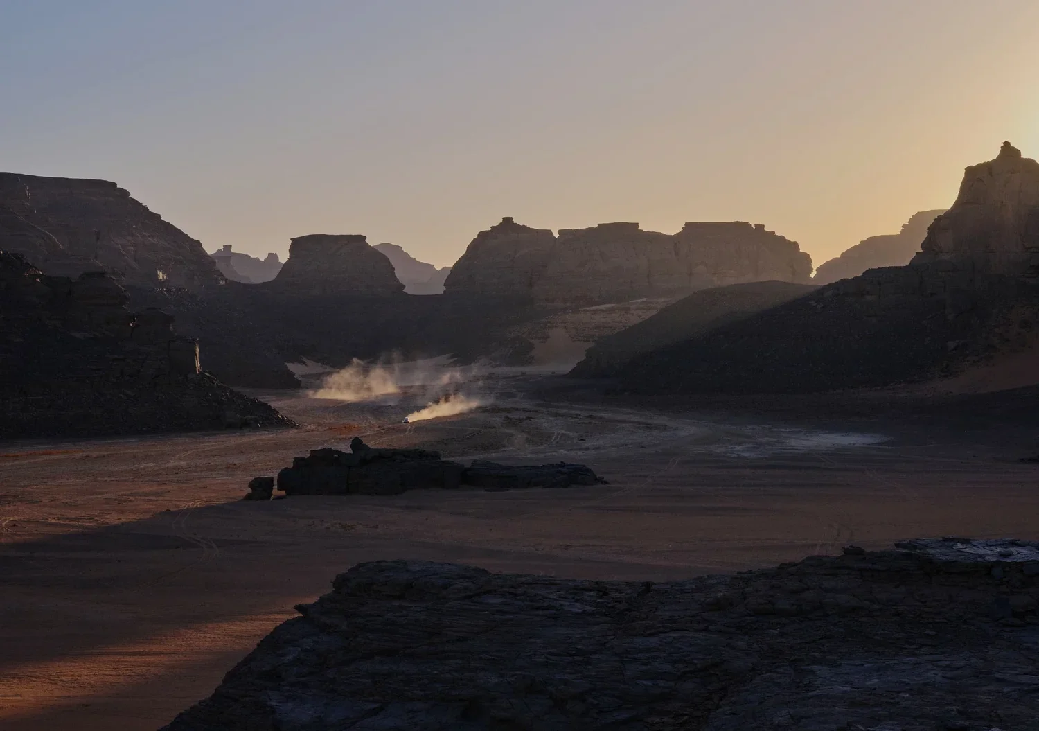 Desert landscape with towering rock formations at sunset or sunrise, with a vehicle kicking up dust in the distance.