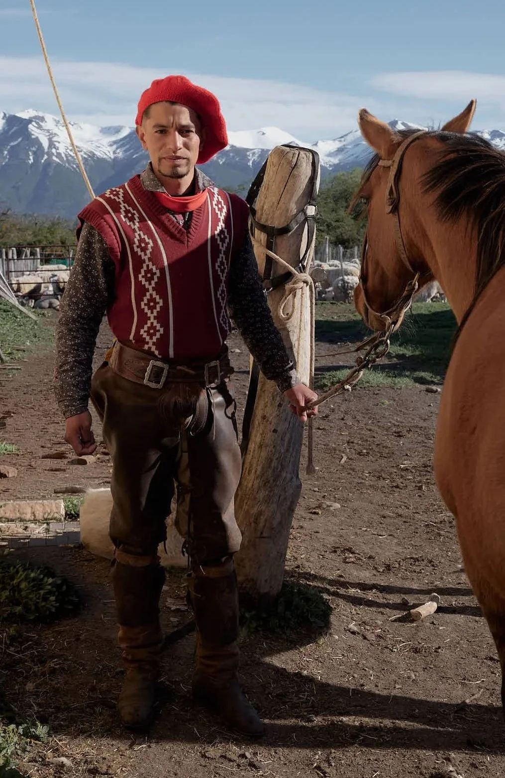 A man dressed in traditional European mountain attire standing next to a brown horse in a rural setting with snow-capped mountains in the background.