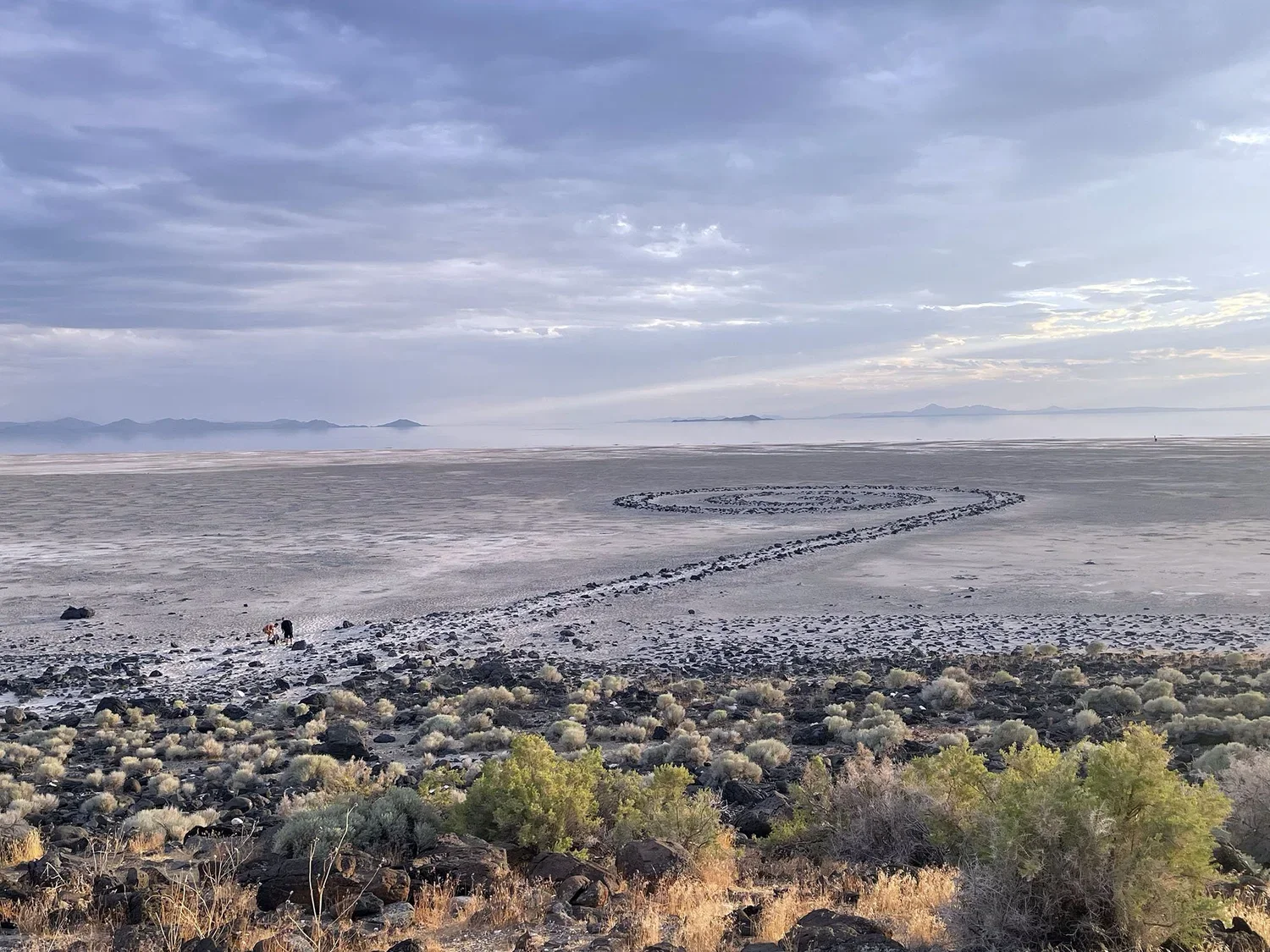 A vast, desolate salt flat landscape with a trail of rocks creating a winding path toward the distant horizon. Sparse vegetation covers the foreground, with small bushes and rocks. The sky is partly cloudy with layers of clouds, and mountains are barely visible in the background.
