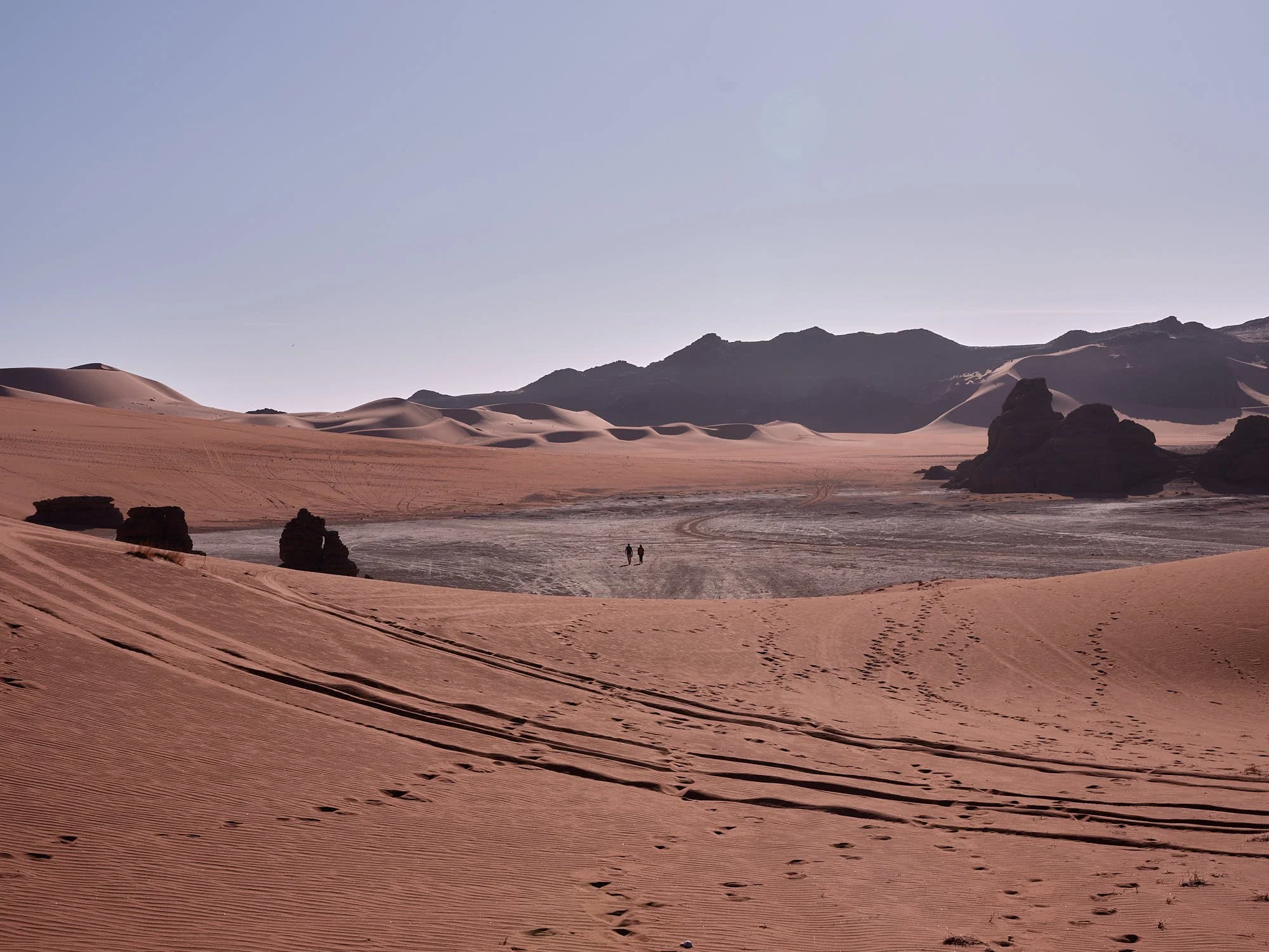 Desert landscape with sand dunes, rock formations, and mountains in the background, under a clear sky.