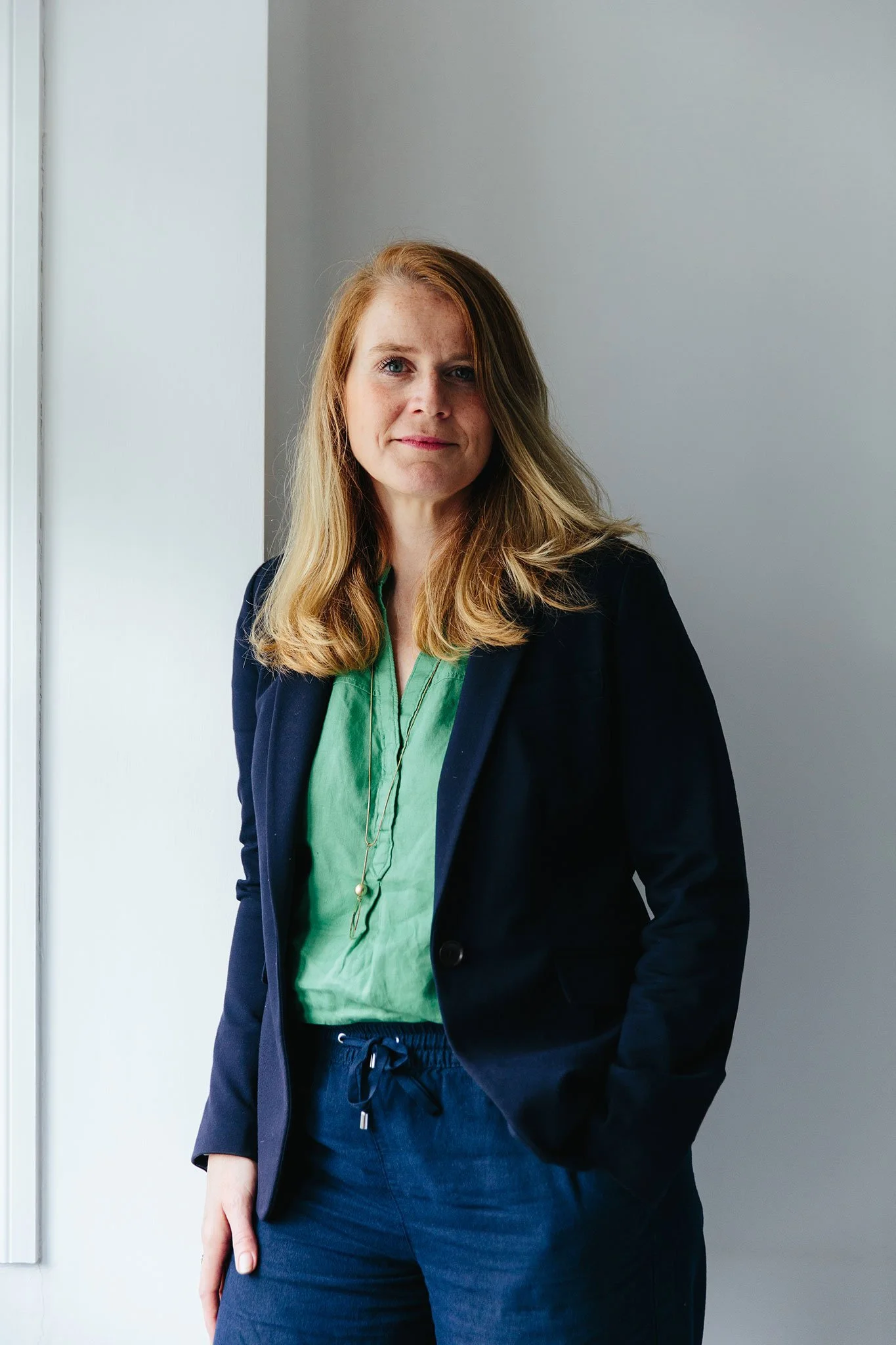 Person in a green blouse and navy blazer standing in a room.