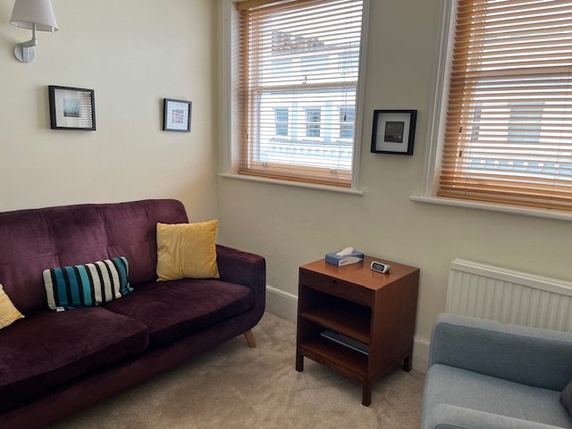 Cozy living room with a purple sofa, striped and yellow cushions, a small wooden side table, blue armchair, framed pictures on the walls, and wooden blinds on the windows.