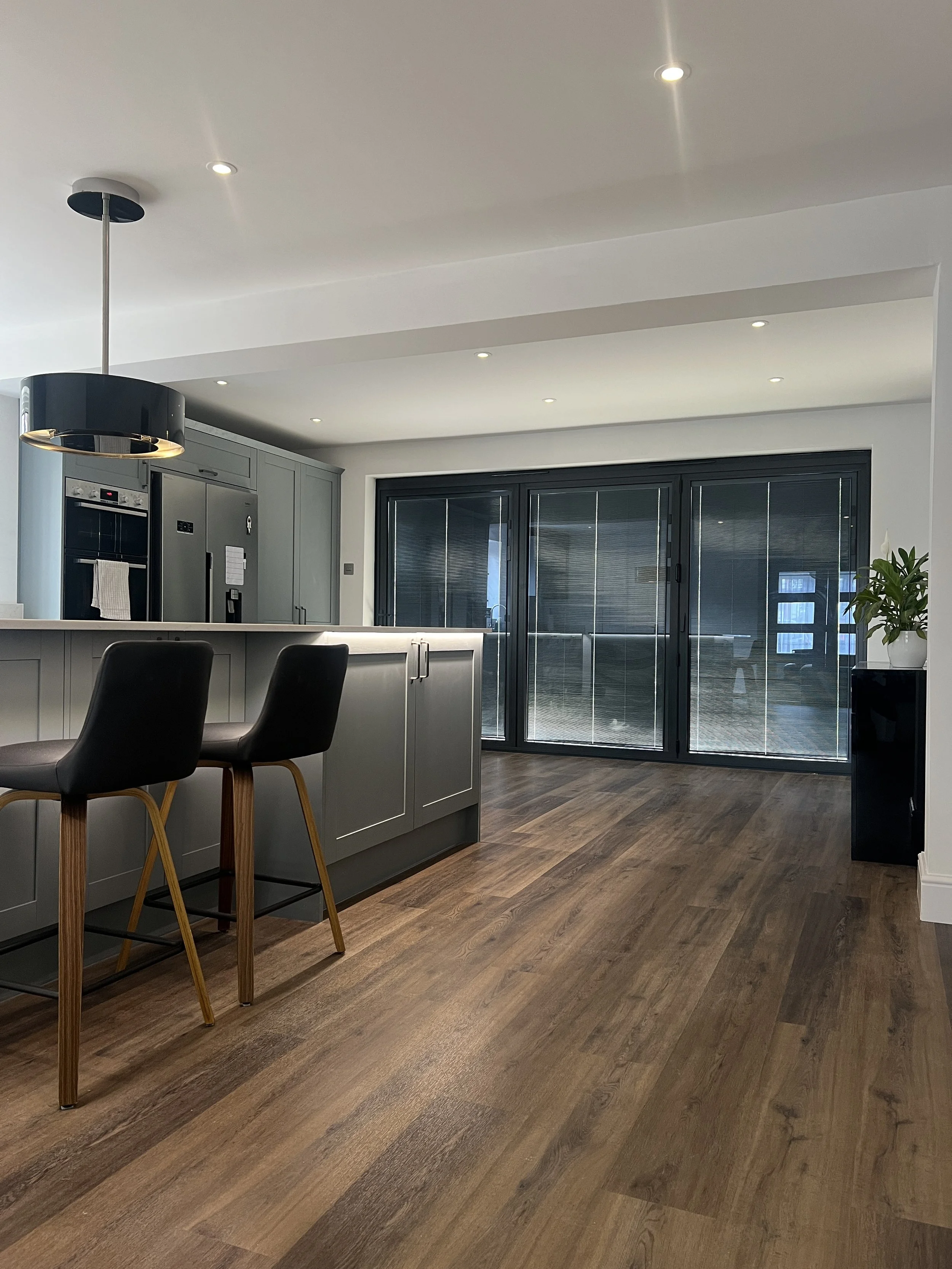 Modern kitchen with dark wood flooring, gray cabinets, black sliding glass doors, black and gold pendant light, and black chairs.