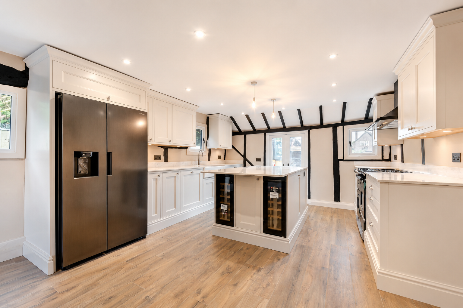 Modern kitchen with black and white cabinetry, wooden floors, stainless steel appliances, including a refrigerator and stove, and a central island with wine storage. Exposed beams on the ceiling and multiple small ceiling lights.