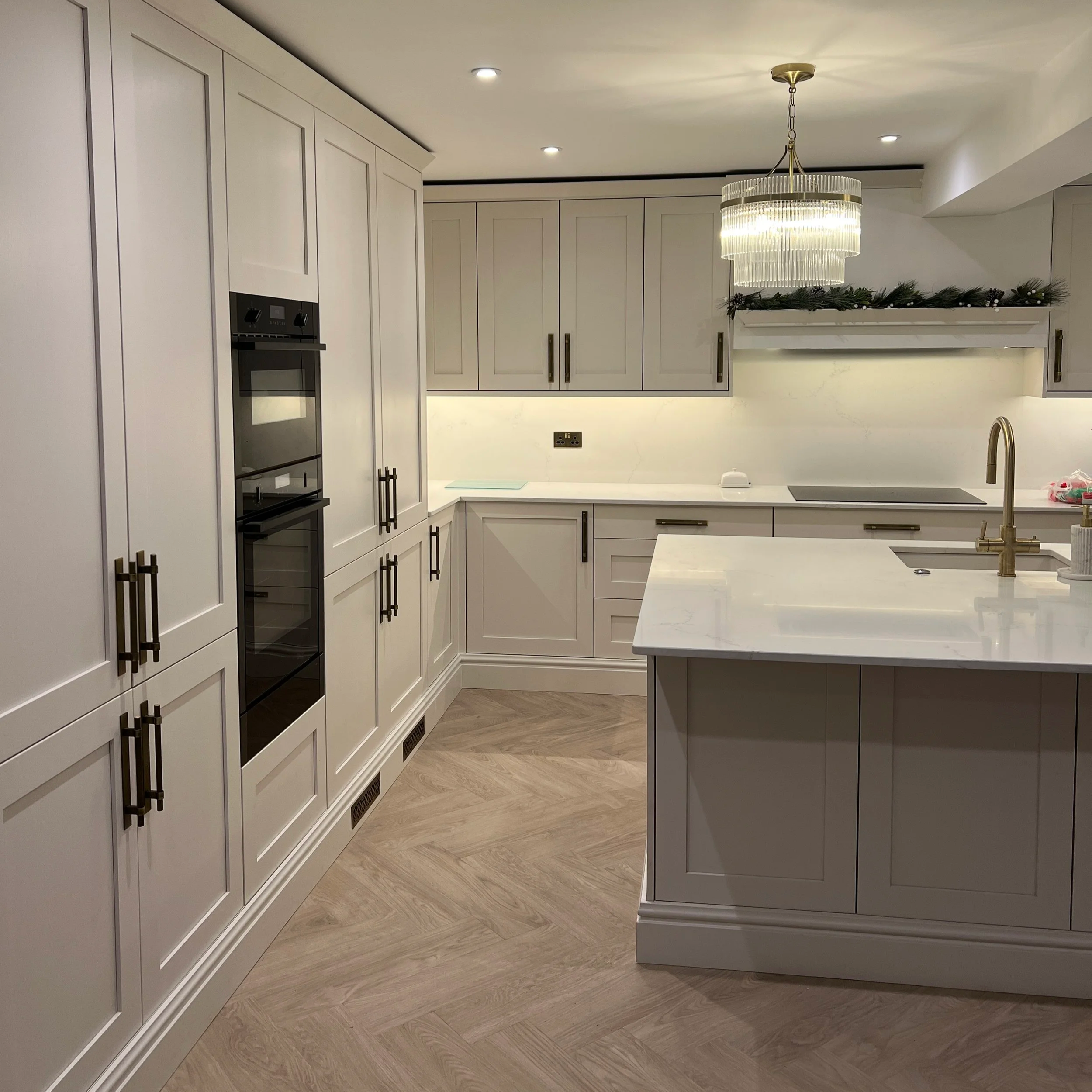 Modern kitchen with white cabinets, a white island, a chandelier, wooden flooring, and Christmas garland above the fireplace