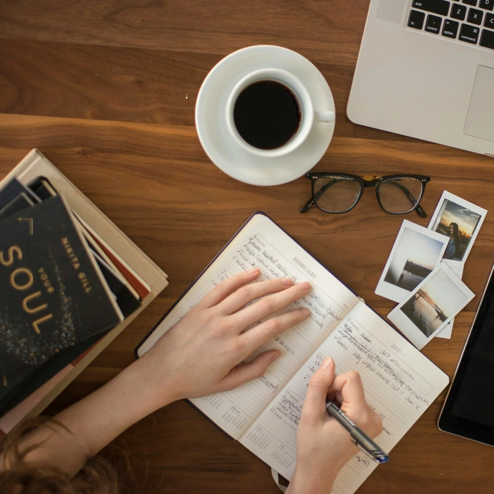 A person writing in a planner on a wooden desk surrounded by a laptop, glasses, a cup of coffee, a pack of books, and several Polaroid photos.
