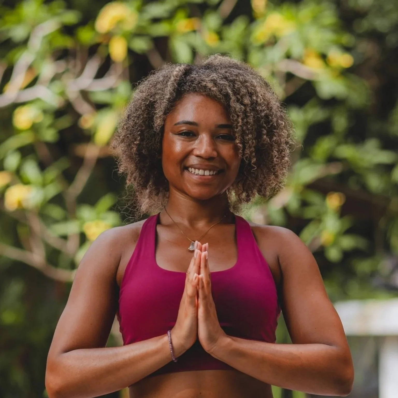 Woman with curly hair smiling outdoors, holding hands in a prayer pose in front of a green leafy background.