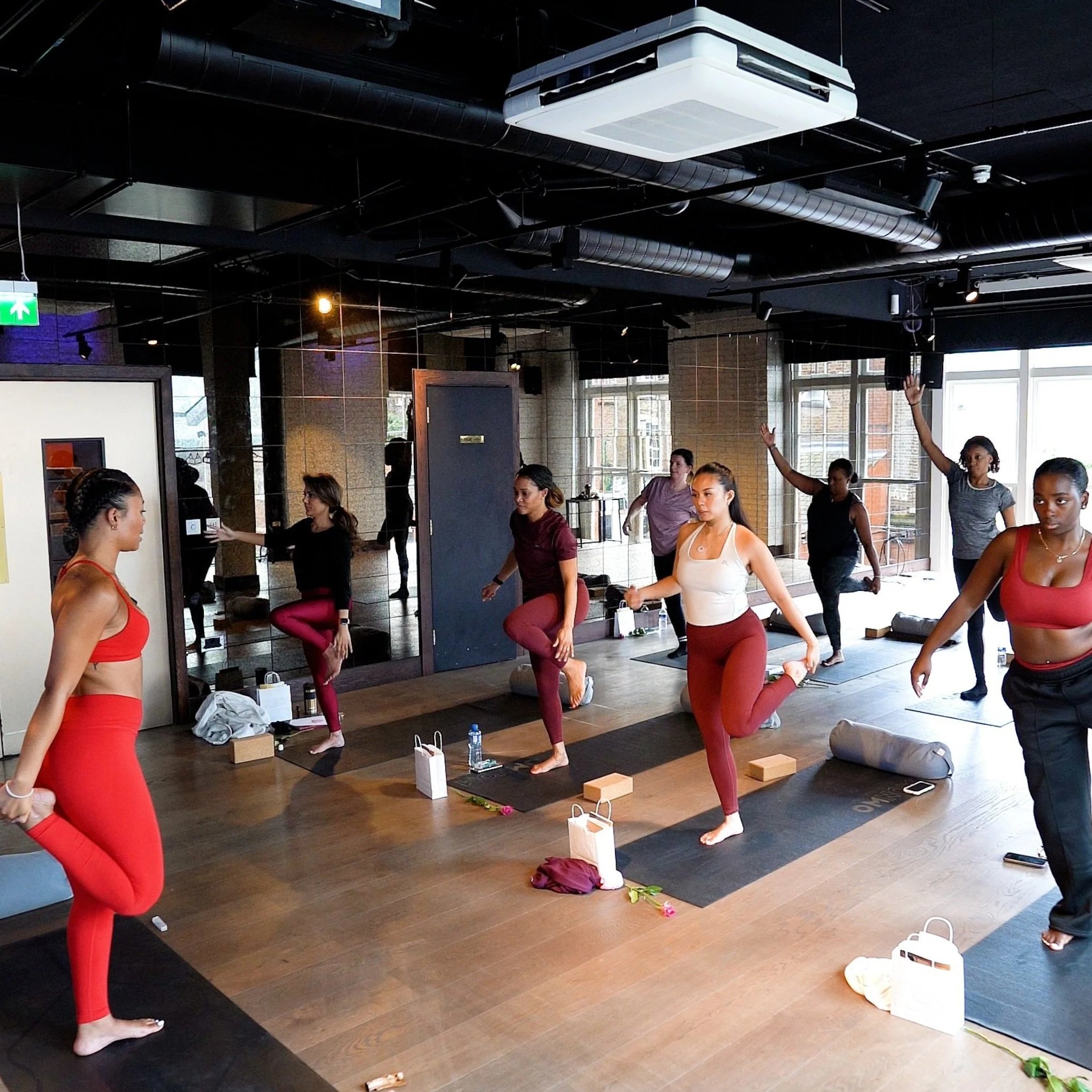 A group of women practicing yoga in a fitness studio, some standing on one leg while holding the other foot behind them, wearing workout clothes, with yoga mats and personal belongings on the floor.