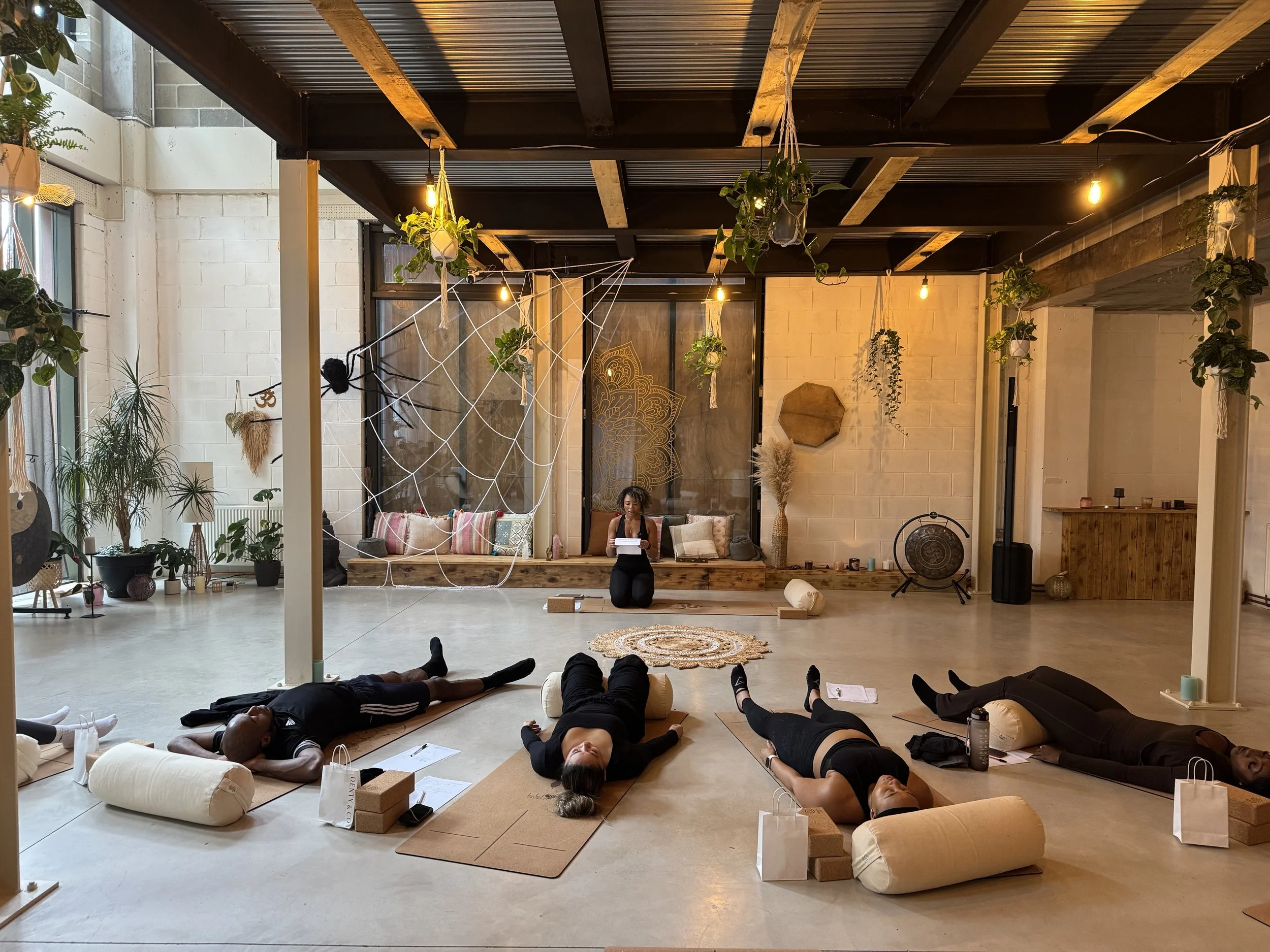 Yoga class in a spacious, decorated studio with participants lying on mats and bolsters, a teacher sitting cross-legged at the front with a laptop, plants, and hanging decor.