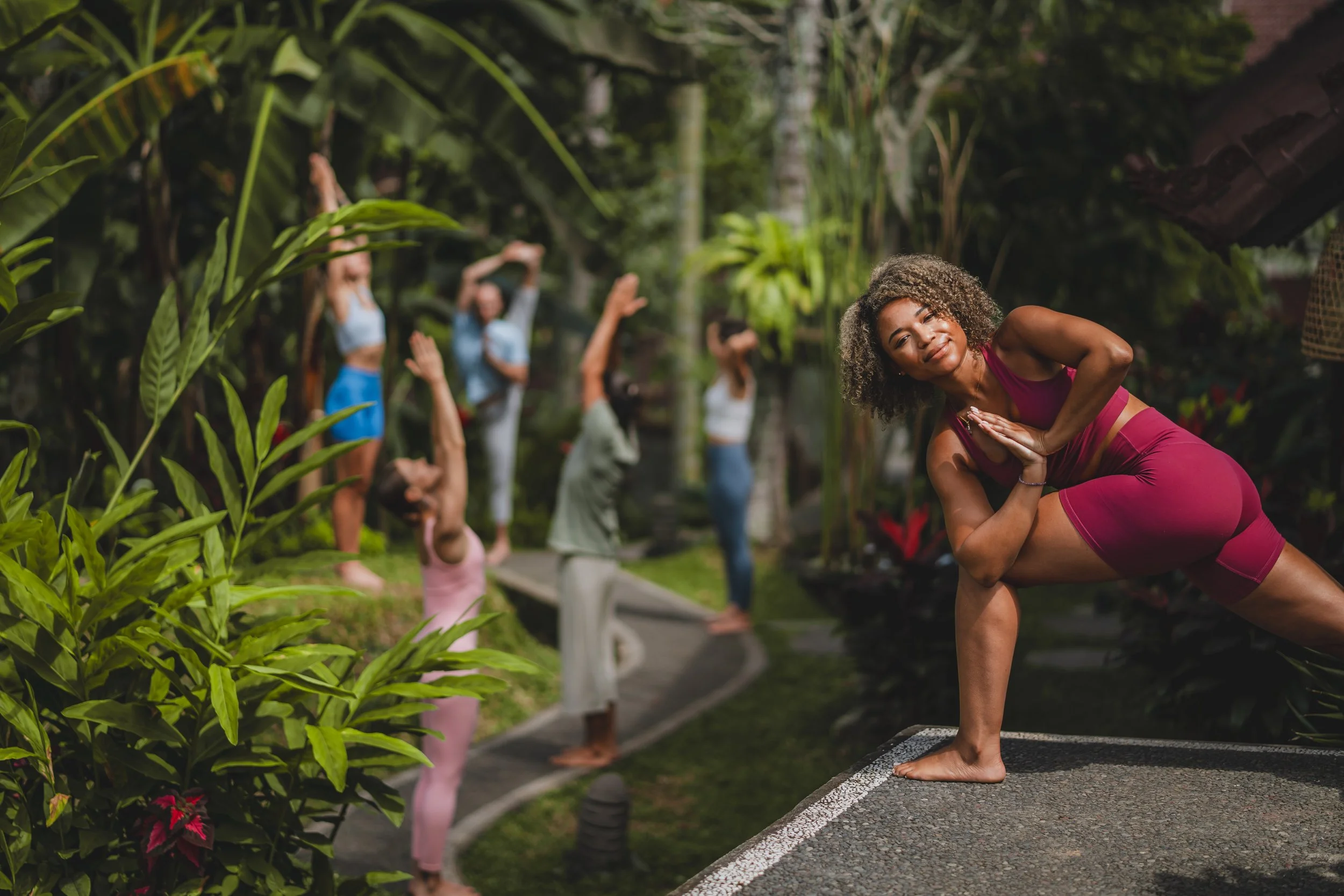 A woman in a pink workout outfit practicing yoga on a platform in a lush, tropical outdoor setting with several other women in the background doing yoga poses.