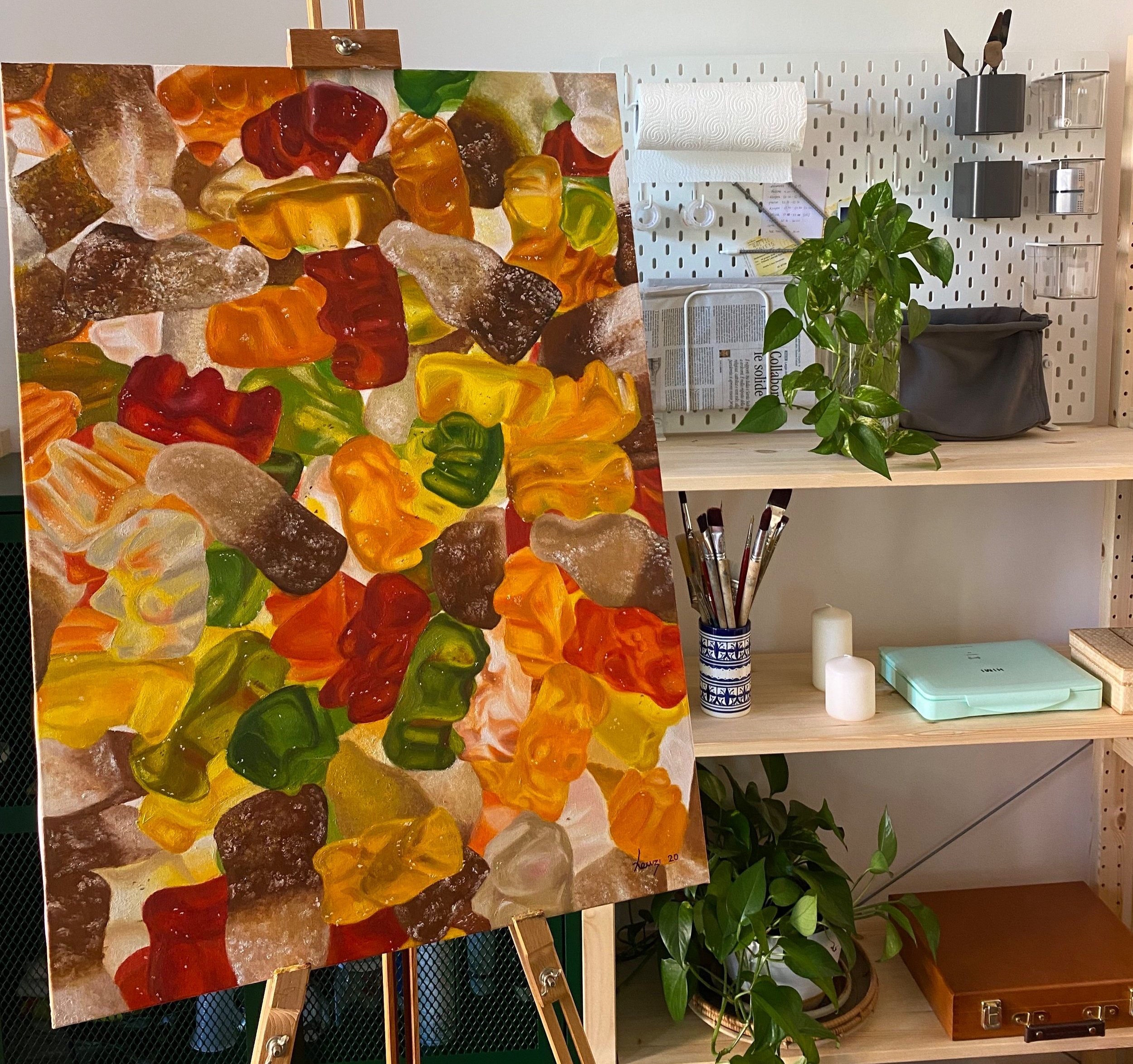 Un quadro con dipinti di caramelle di varie forme e colori, posato su un cavalletto. Sullo sfondo si vede una parete con alcuni scaffali con contenitori e piante verdi.