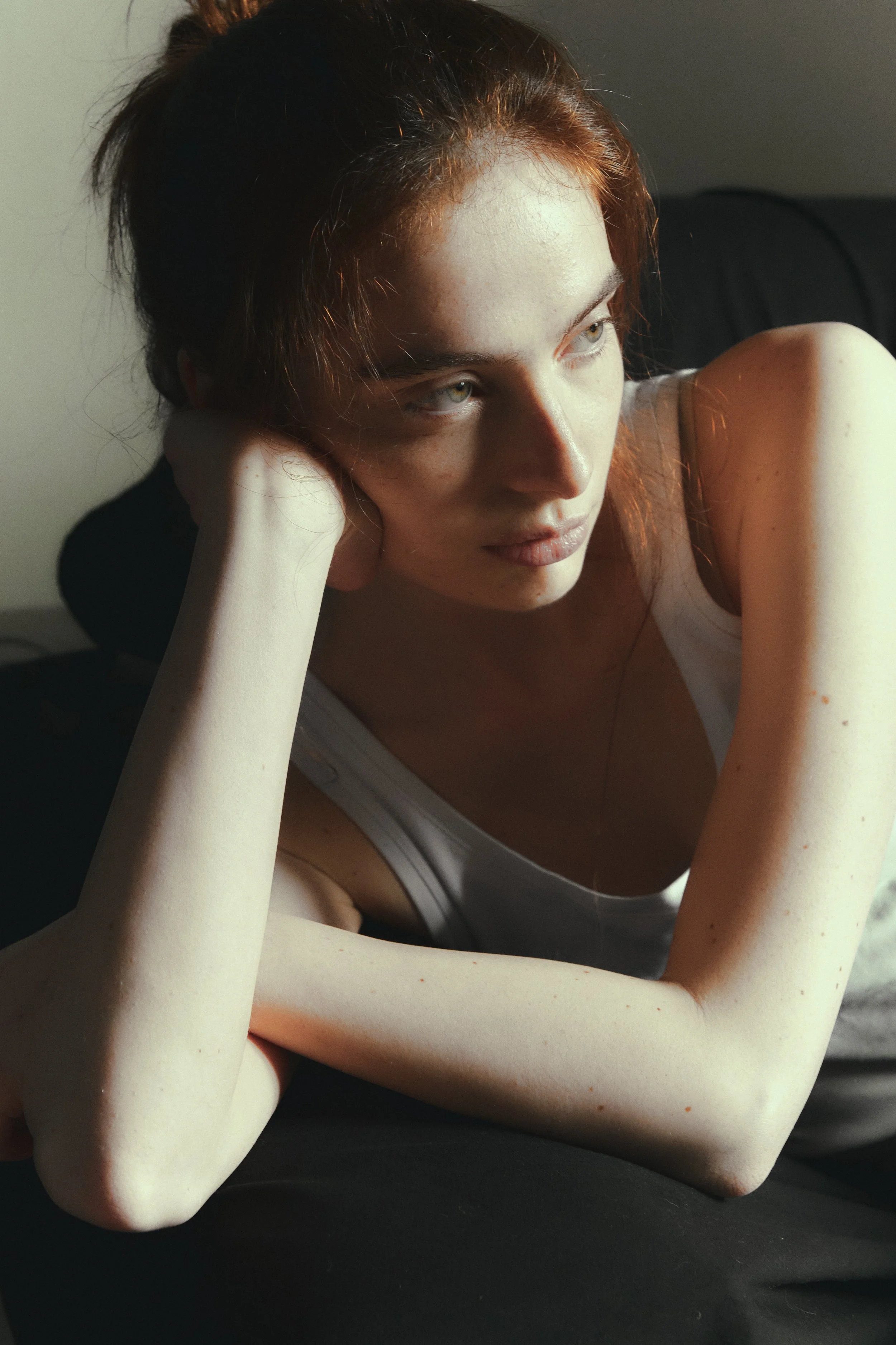Ragazza con capelli rossi seduta con il viso appoggiato sulla mano, in luce naturale, con braccio e spalla visibili, indossa una canottiera bianca.