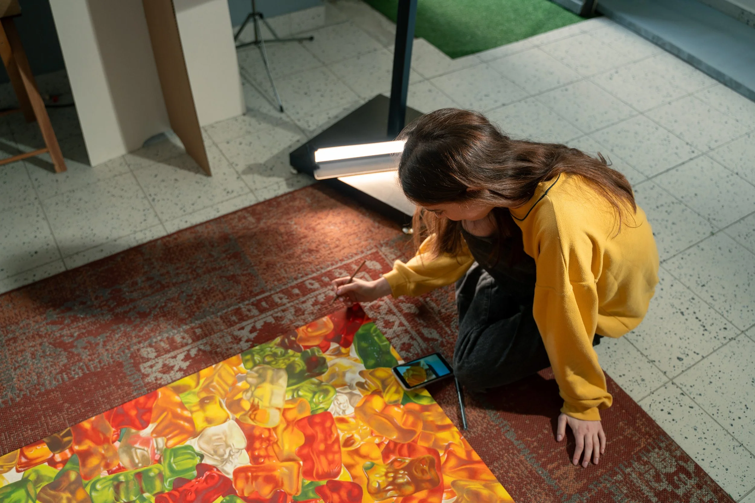 Persona seduta sul tappeto che riprende un'immagine da uno smartphone, con un quadro astratto di colori caldi sul pavimento davanti a lei.