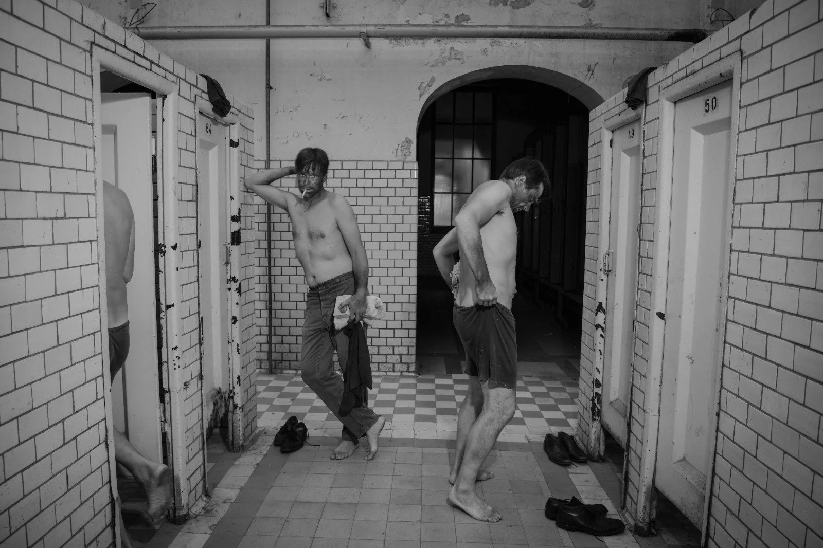 Two shirtless men in a locker room getting dressed, with one adjusting his pants and the other holding a towel and cigarette, among open locker doors and shoes on the floor.