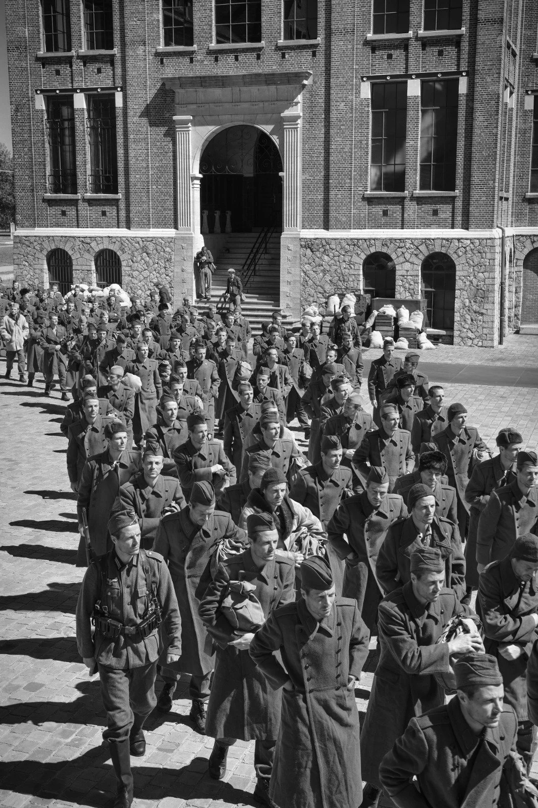 Black and white photo of a large group of soldiers marching in formation outside a brick building with steps and large windows.