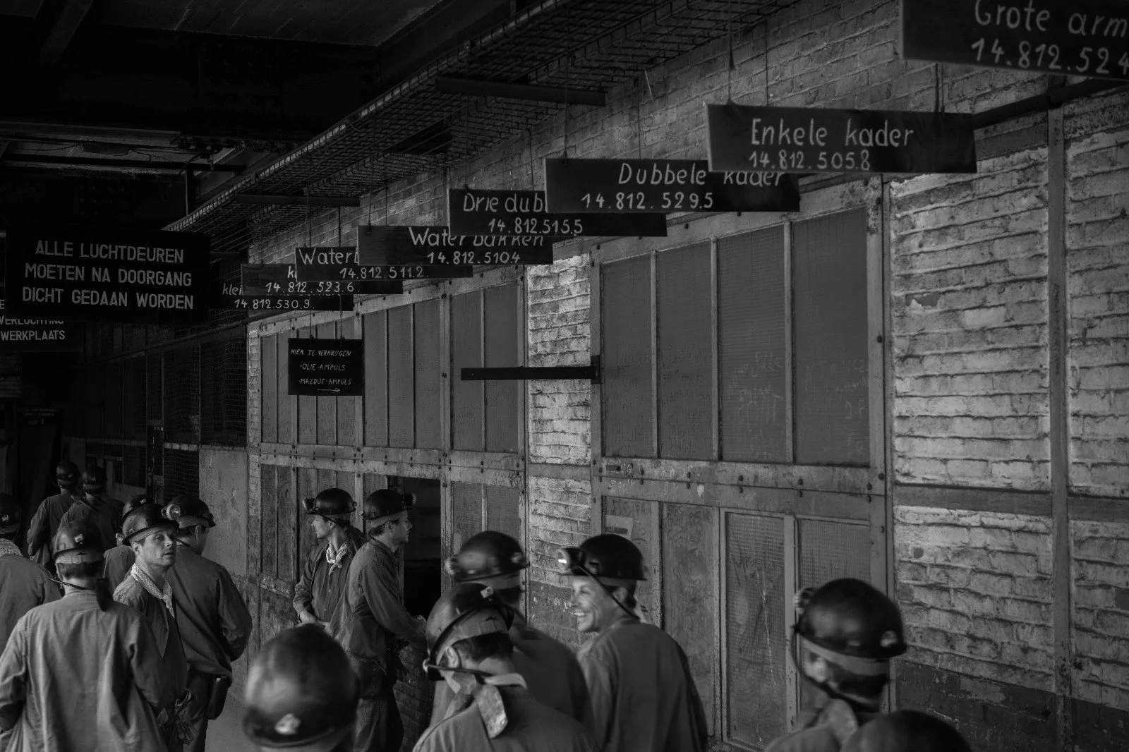 A group of factory workers wearing safety helmets and masks walking and standing in a warehouse with brick walls. Above them, signs in Dutch display different types of stock and their codes, along with a large sign on the left that states: