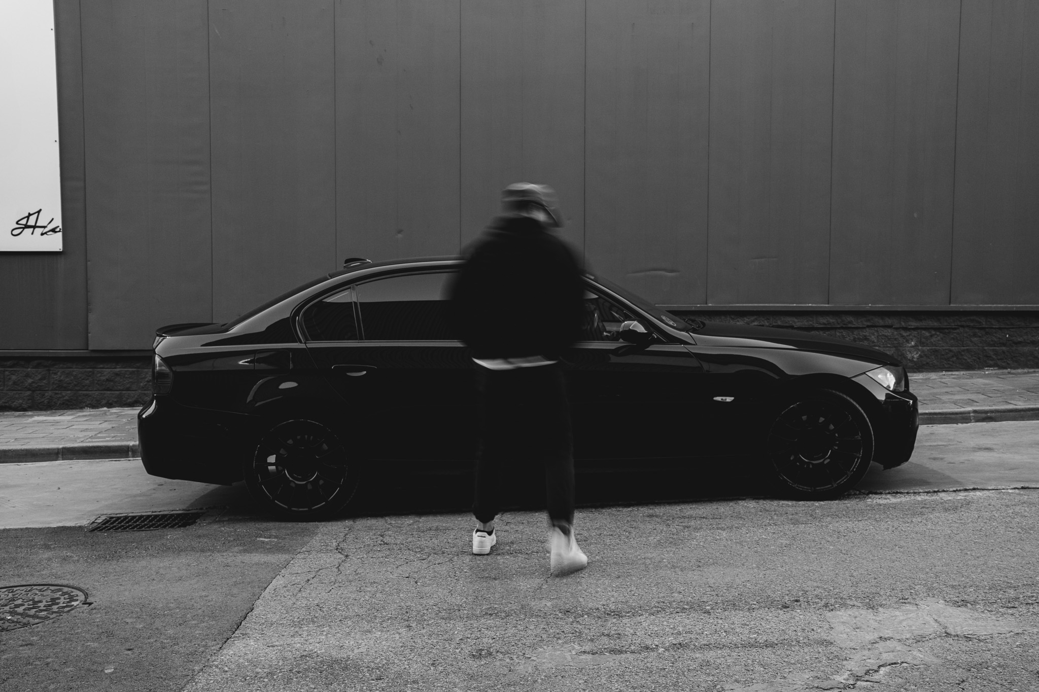 Una persona con casco caminando frente a un coche negro estacionado en la calle, en un entorno urbano con muro y acera.