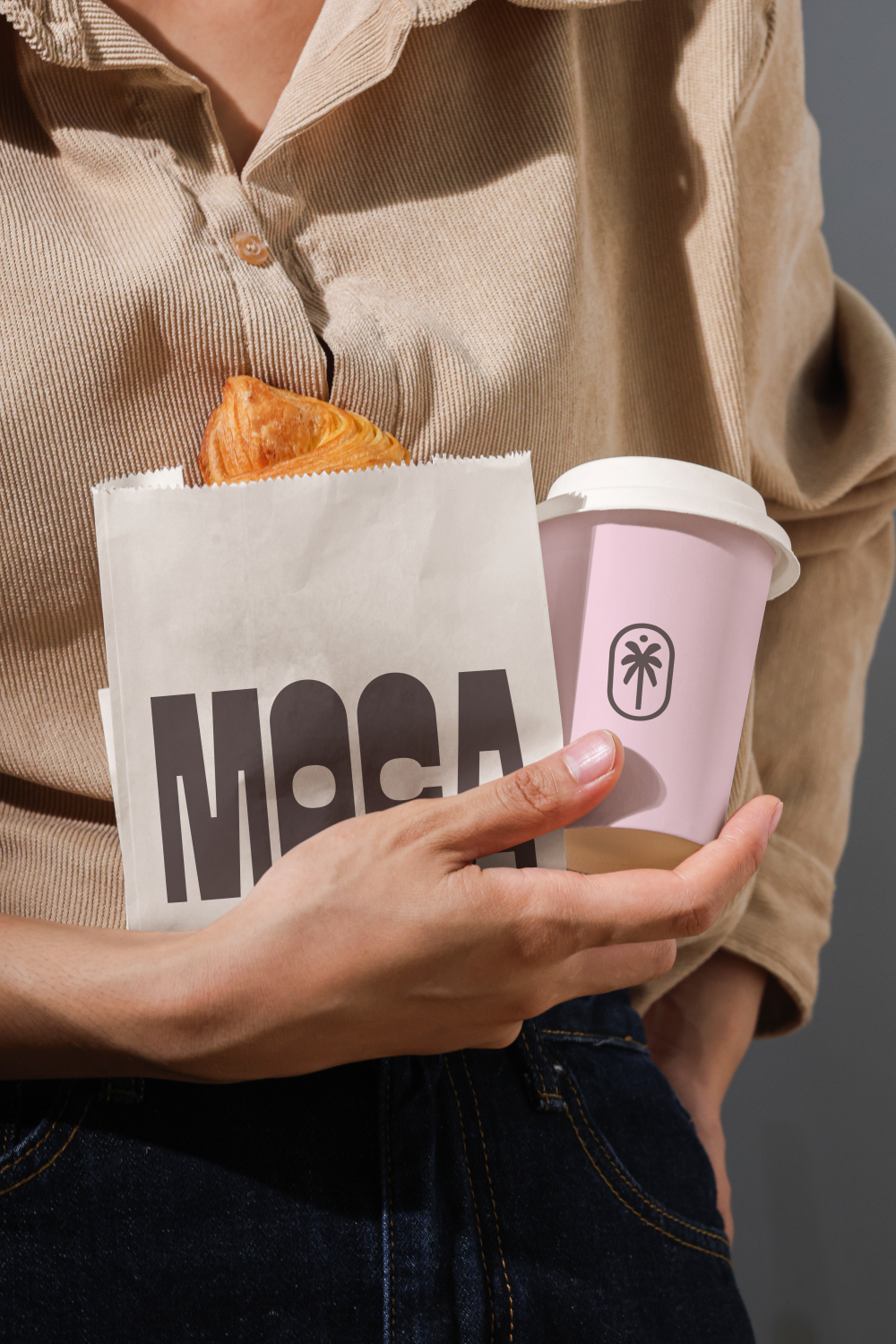 Persona sosteniendo una taza rosa, una bolsa de papel con un croissant y un vaso de café con tapa blanca.