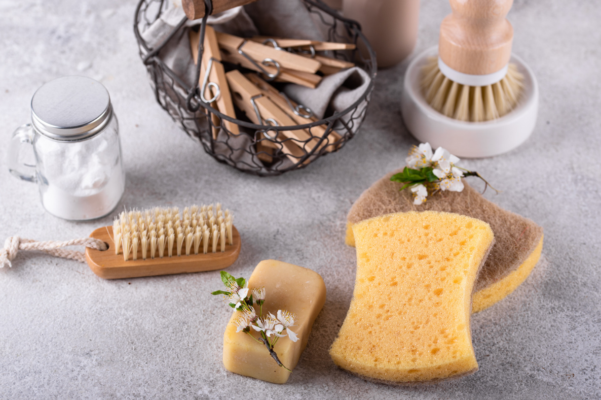 Cleaning supplies on a gray surface, including a jar of baking soda, natural bristle brushes, sponges, wooden clothespins in a wire basket, and a bar of soap with flowers.
