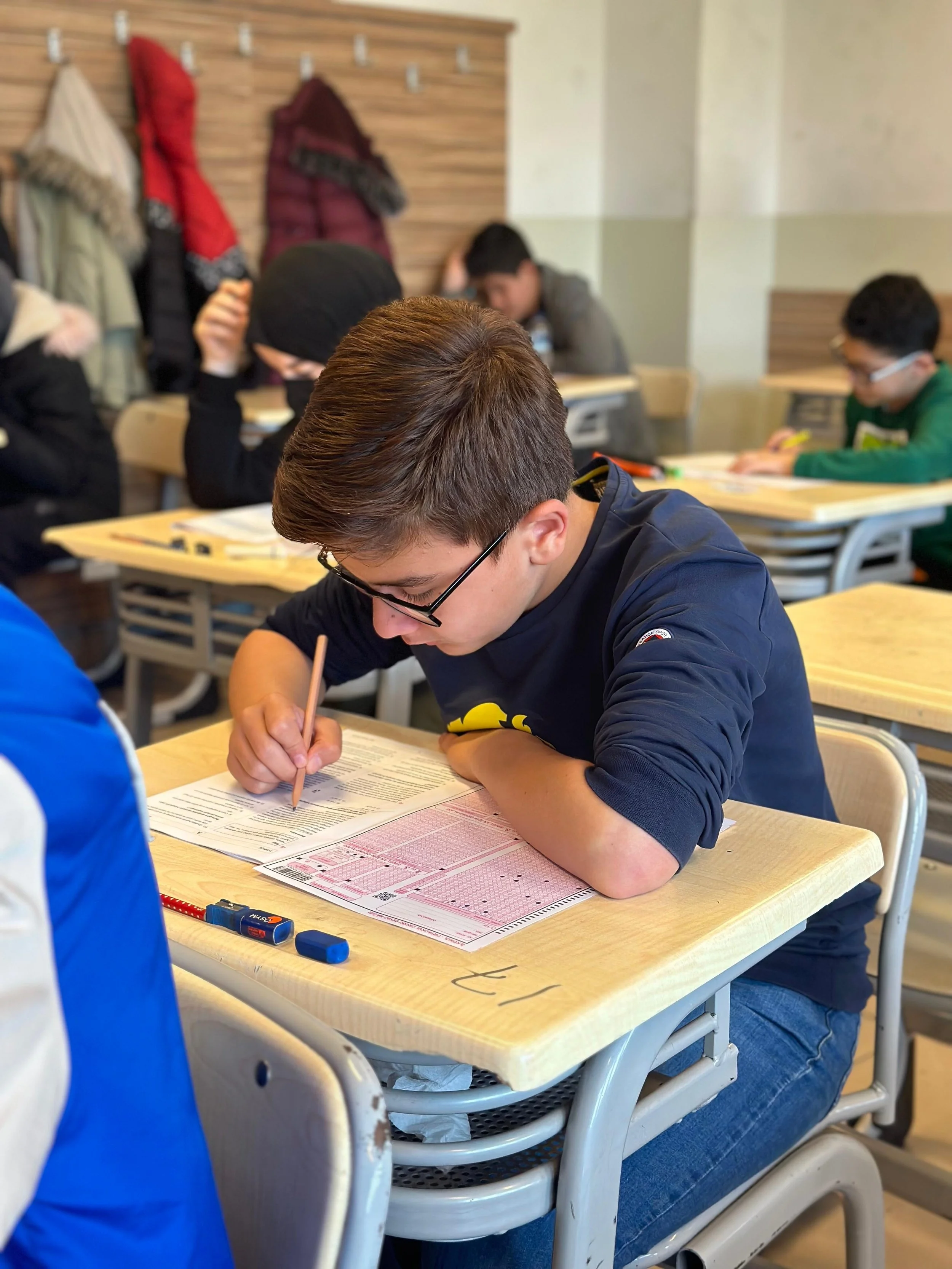 Sınav yapan bir öğrenci sınava odaklanmış ve kalemle cevap işaretleri yapıyor; arka planda diğer öğrenciler de sınava devam ediyor.