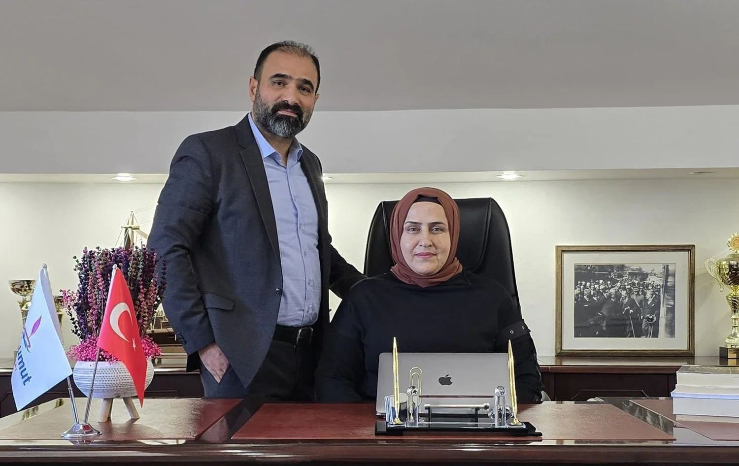 İki kişi, ofis ortamında, bir kadın ve bir erkek. Kadın, ofis sandalyesinde, dizüstü bilgisayar ve birkaç resmi belgeyle çalışma masasının arkasında oturuyor. Erkek ayakta duruyor, kadın ise sandalyesinde. Ofis dekorunda Türk bayrağı ve başka bir bayrak, çiçekler, tablolar ve ödüller bulunuyor.