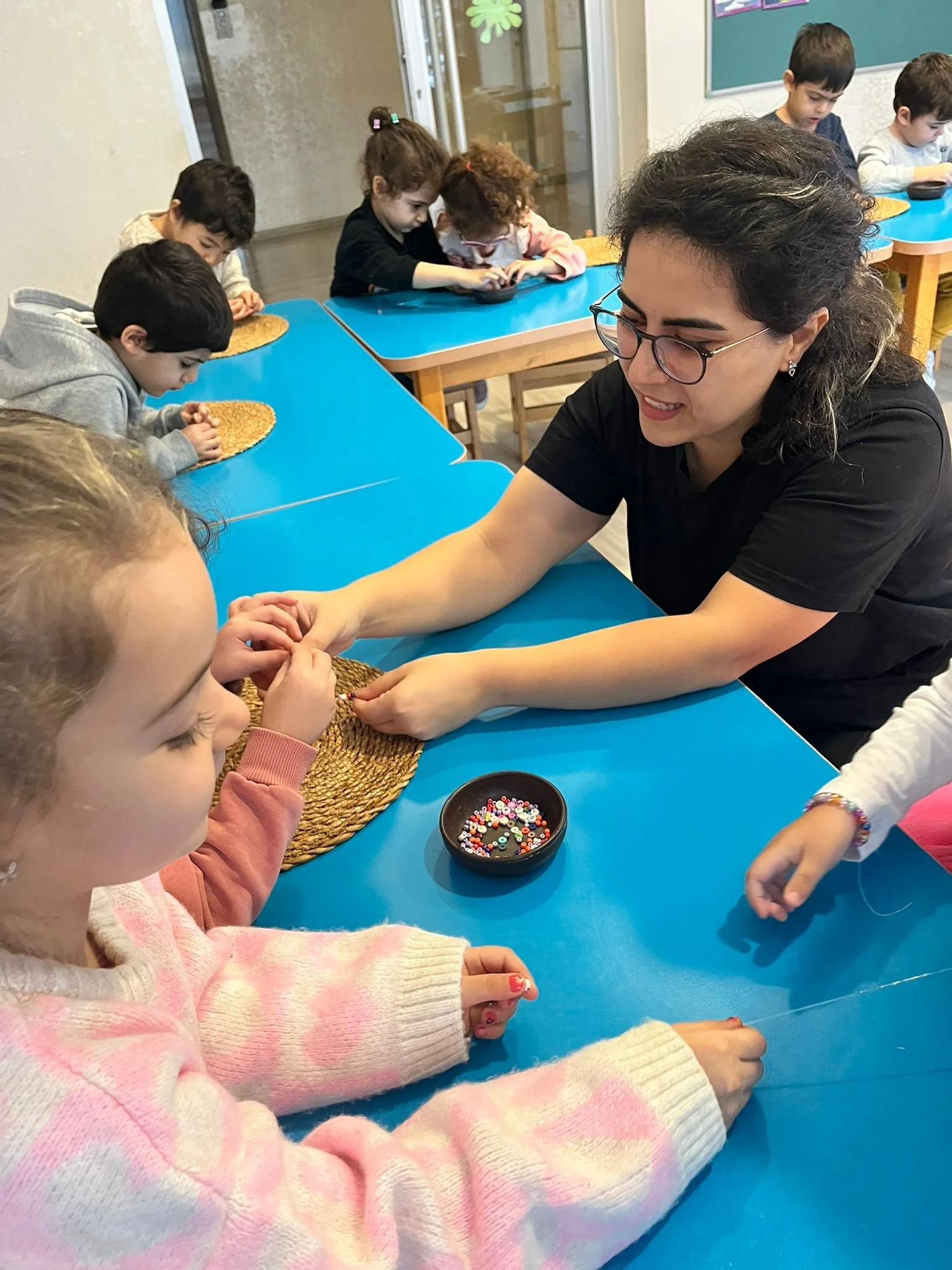Bir öğretmen ve çocuklar boncuk dizme etkinliği yapıyor, masa mavi ve boncuklar siyah bir kapta bulunuyor.