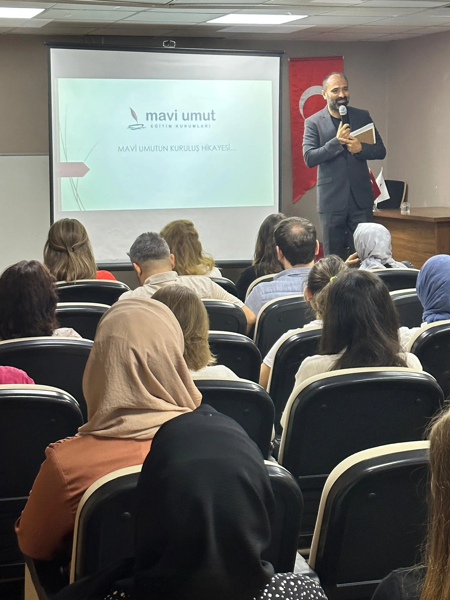 Bir konferans veya seminer salonunda bir adam mikrofon tutarken sunum yapıyor. Sunum ekranında 'mavi umut' ve 'MAVİ UMUTUN KURULUŞ HİKAYESİ...' yazıyor. Arka planda Türk bayrağı askıya alınmış.