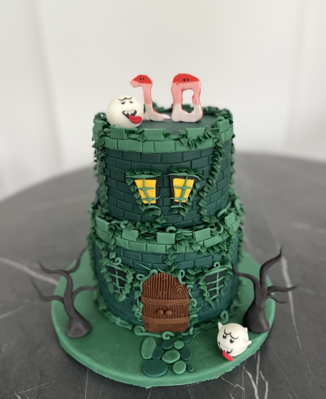 A multi-tiered fantasy castle cake decorated with green bricks, yellow windows, a wooden door, vines, and leaf details. It features ghost and flamingo figures on top, with ghost faces on the base. The cake is on a green base with trees and stones.