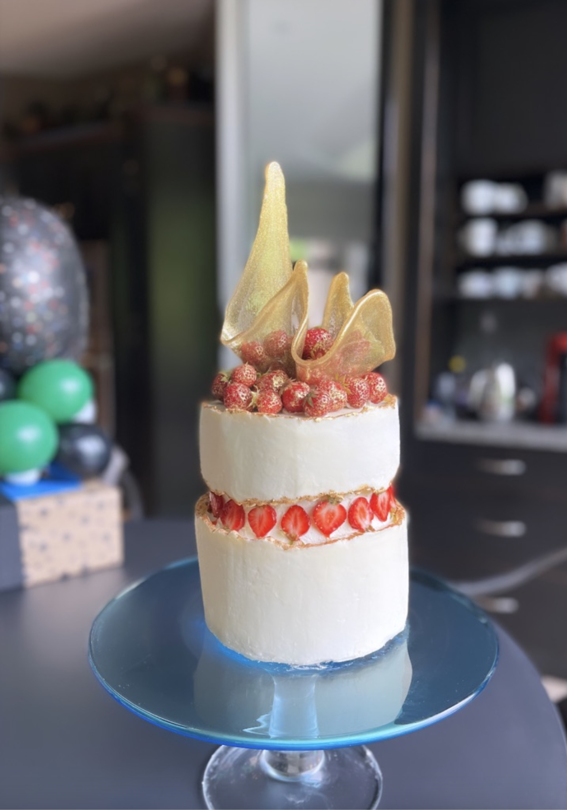 Two-layer white frosted cake with sliced strawberries around the middle, topped with red berries, and decorated with gold-tinted sugar sculptue on top, served on a blue cake stand.