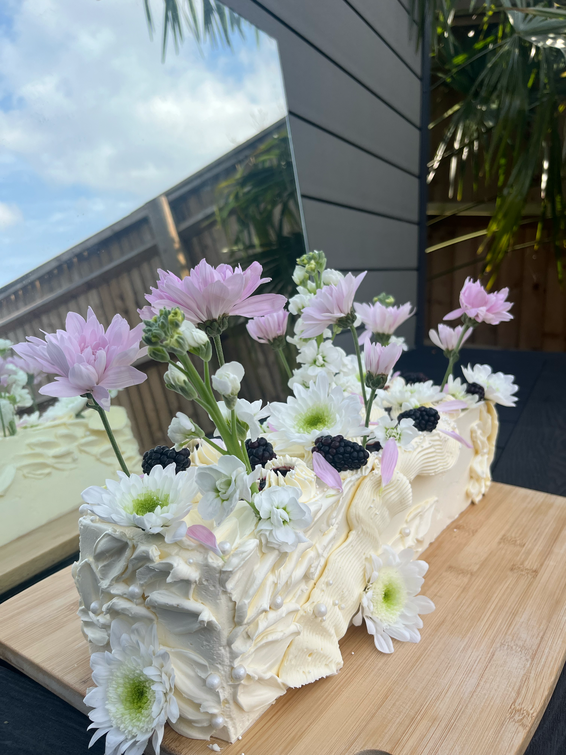Cream-colored cake decorated with white and pink flowers, blackberries, and including a mirror reflecting the sky and garden background.