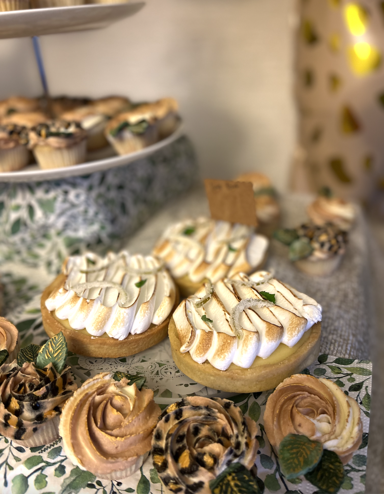 Assorted desserts on a decorative cake stand and platter, including lemon meringue tarts with toasted meringue and slices of rose-shaped cake with swirls of caramel and chocolate, garnished with small leaves.