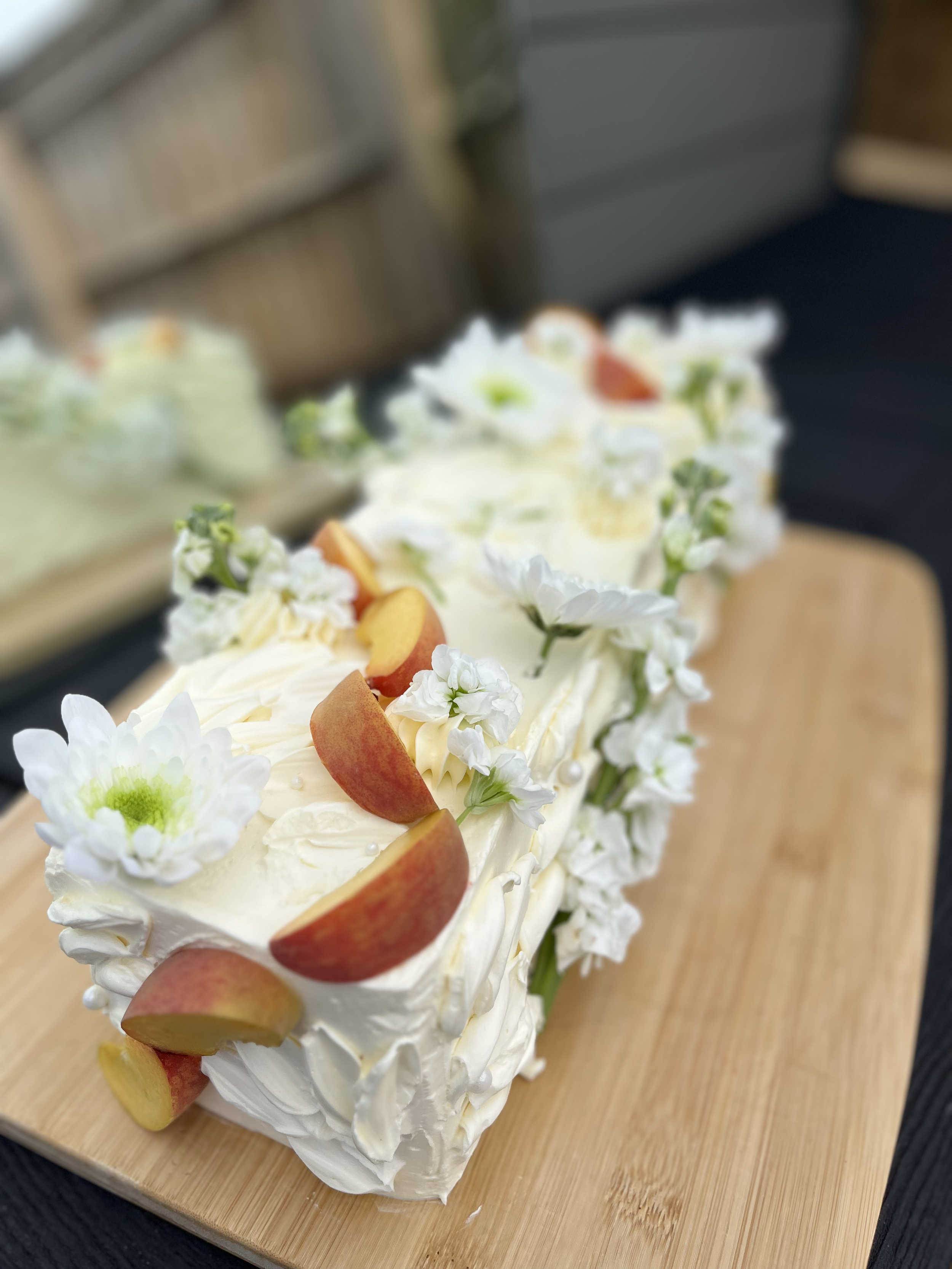 A white frosted cake decorated with slices of peaches and white flowers, placed on a wooden board.