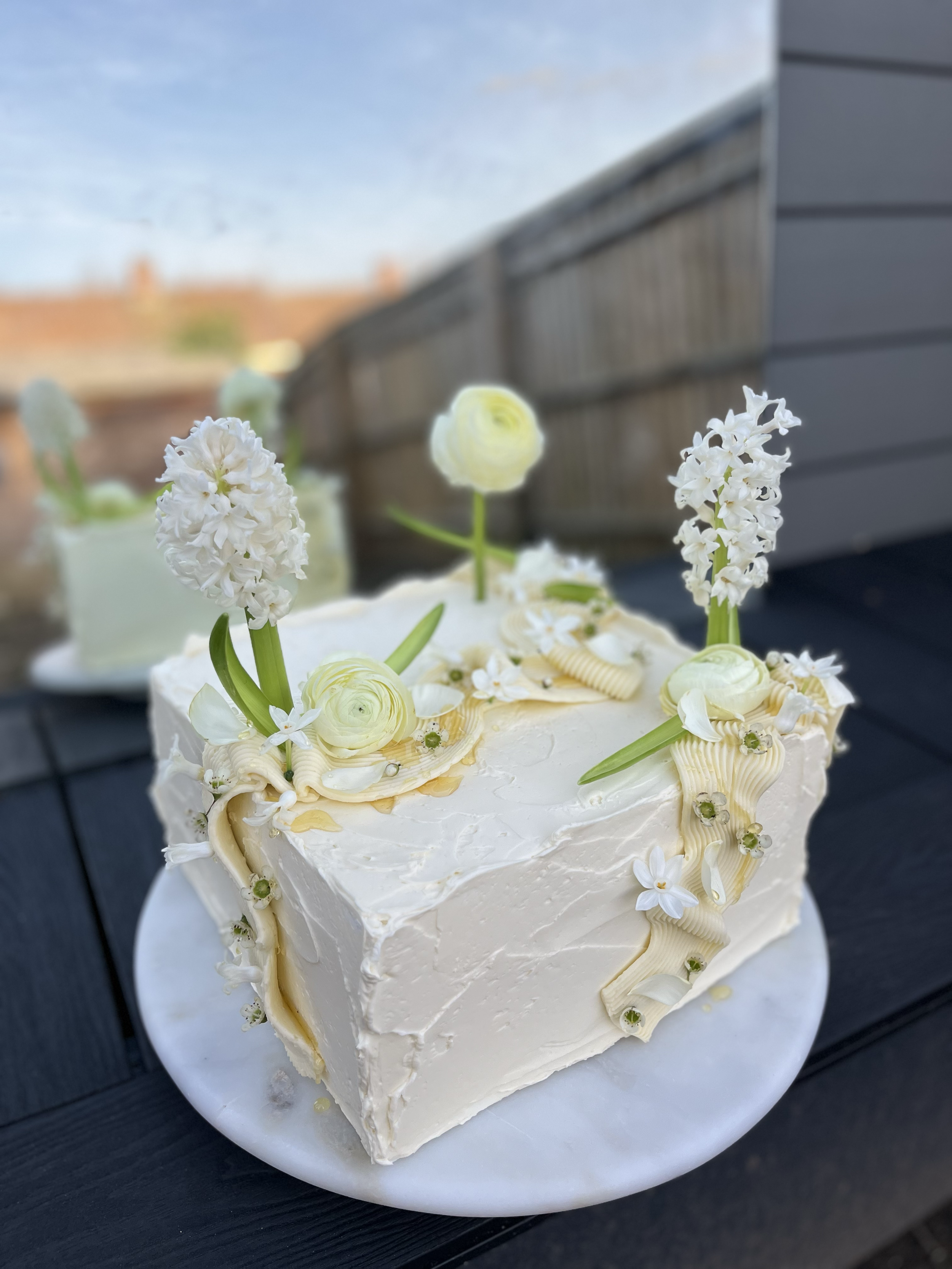 Square white wedding cake decorated with white flowers, green leaves, and cream-colored icing accents, placed on a white marble cake stand outdoors.