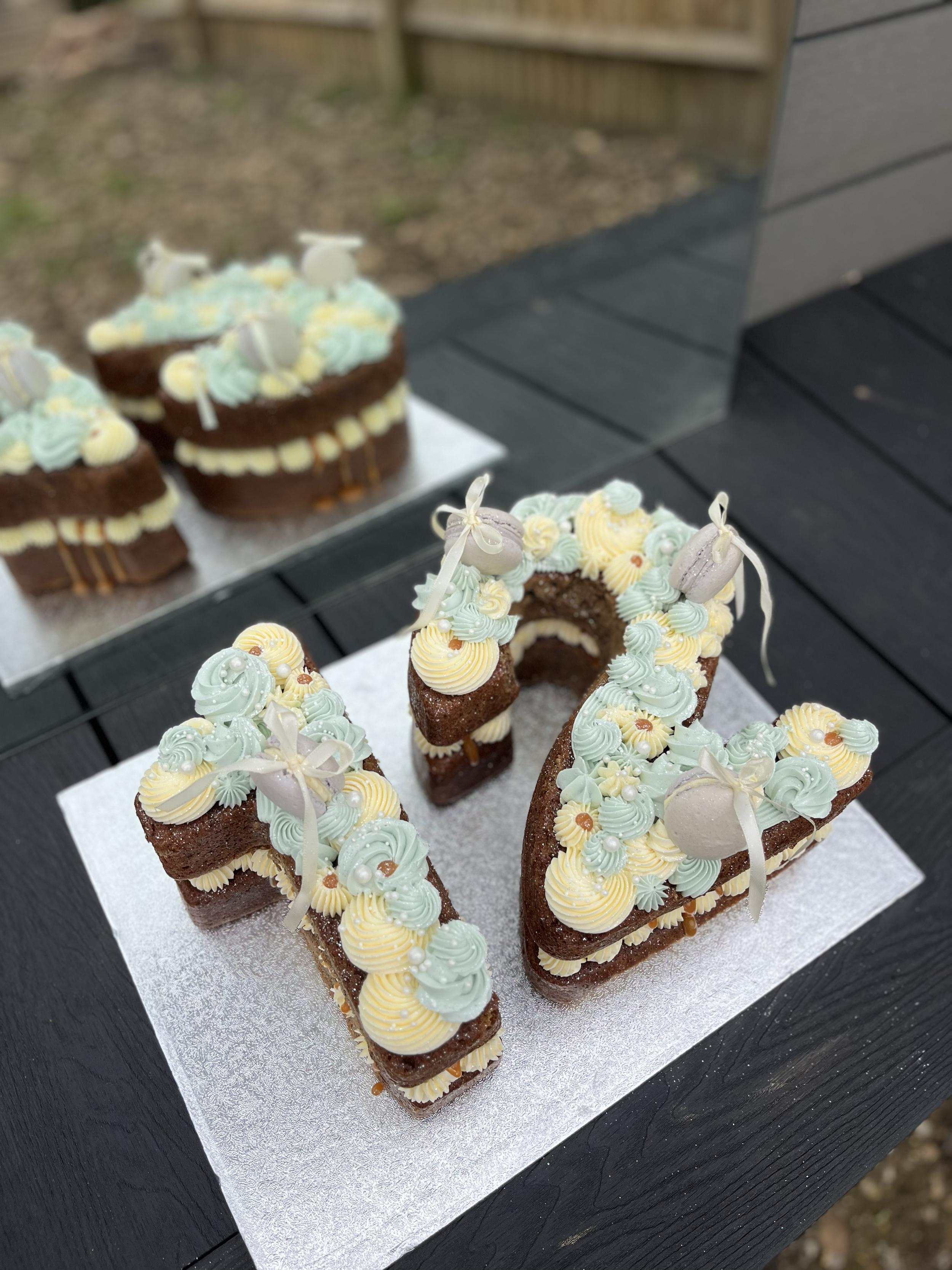 Cake shaped like the numbers 17, decorated with pastel icing, small candies, and white ribbons, on a silver board on a black table outdoors.