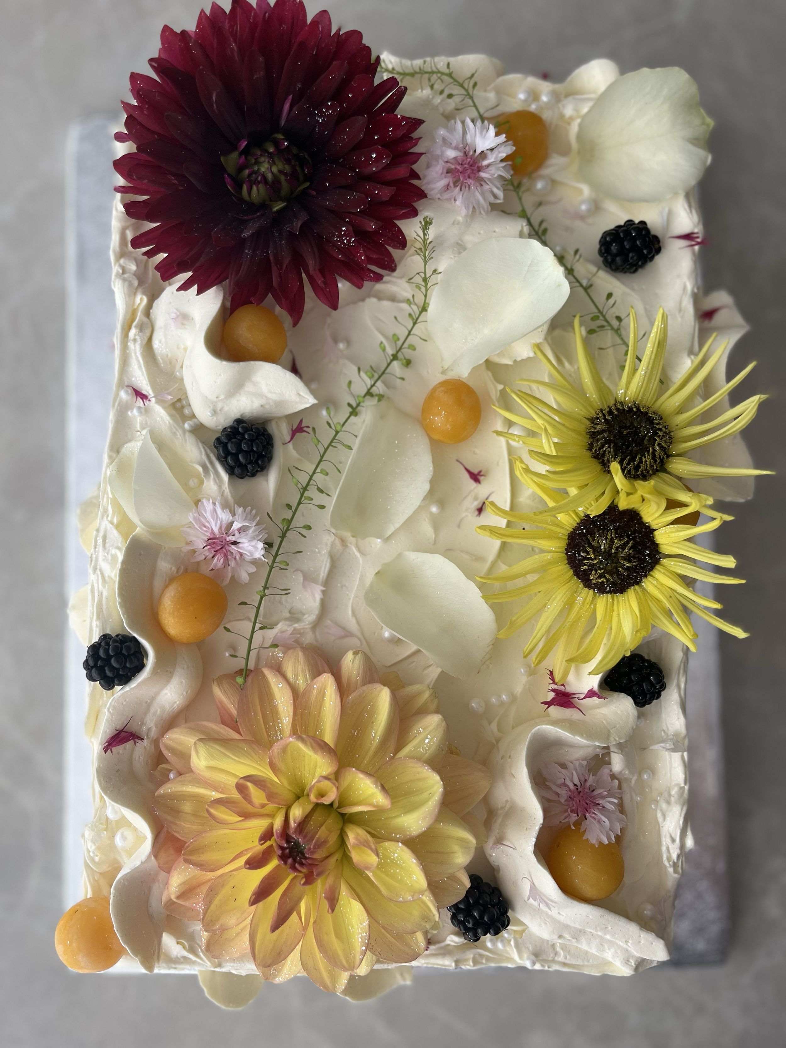 Decorated cake with edible flowers, white chocolate petals, blackberries, yellow and orange berries, and small pink flowers.