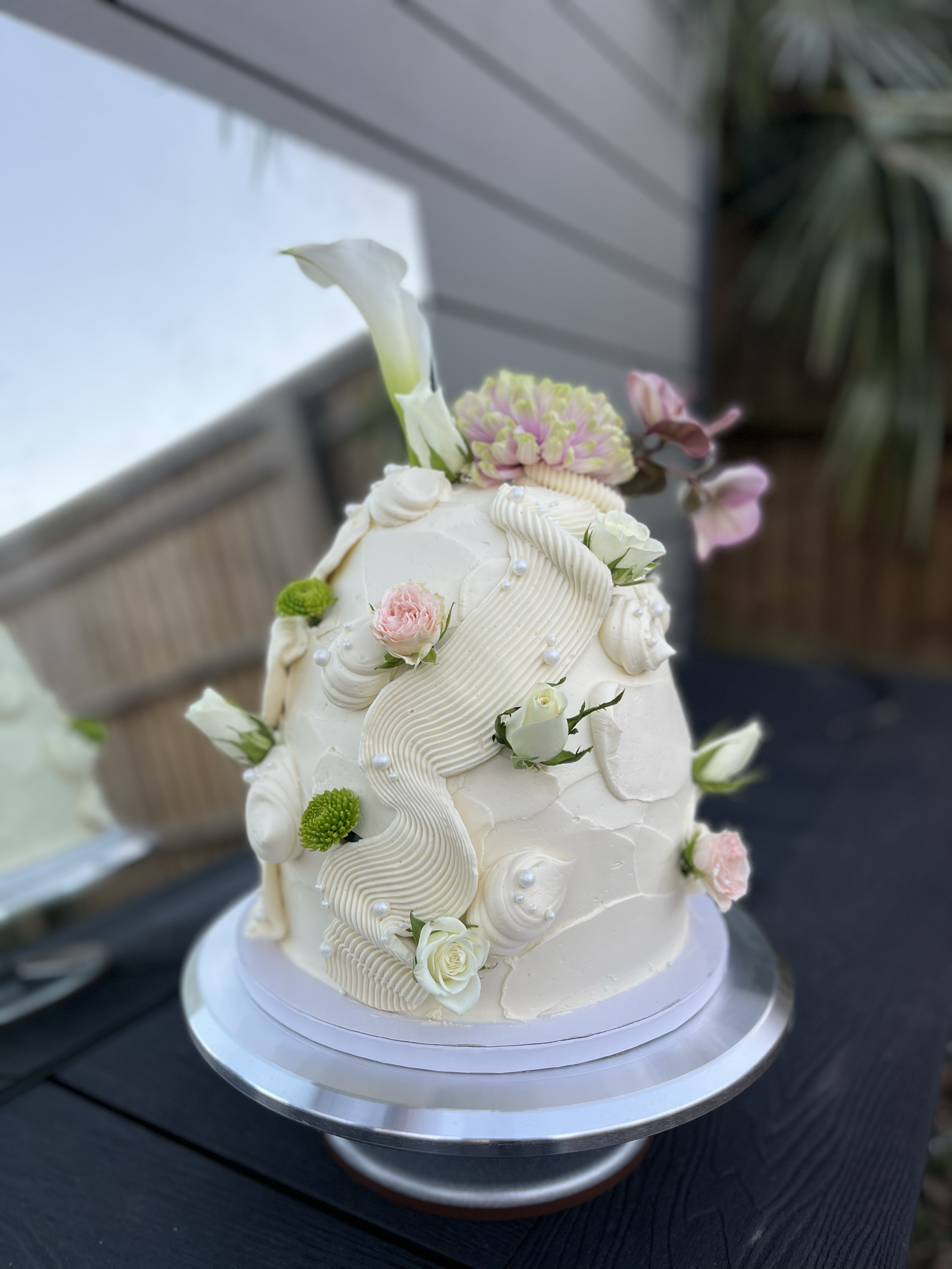 A decorated white wedding cake with floral accents and piped icing, placed on a silver cake stand outdoors.