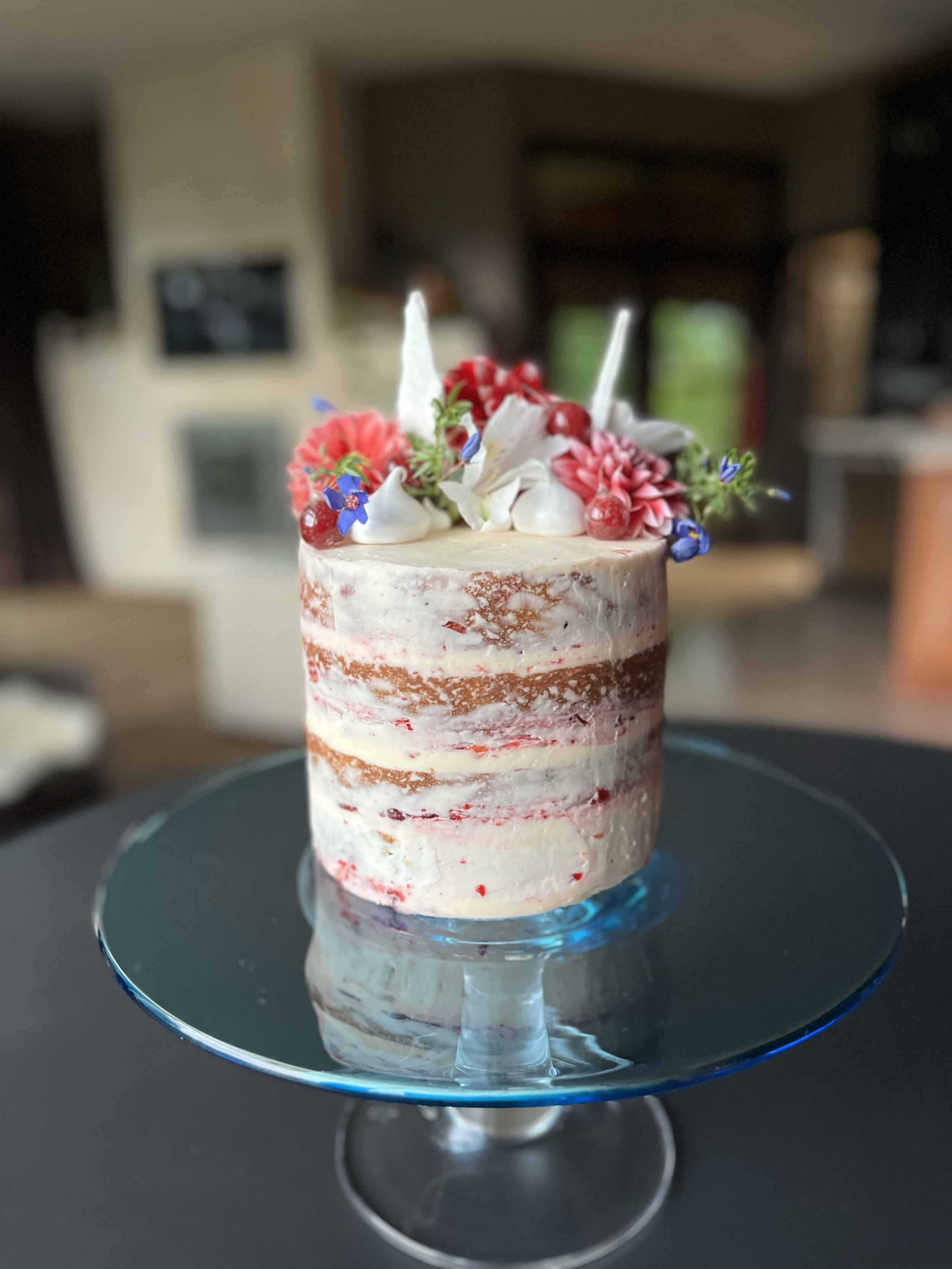 A partially decorated layered cake with white icing, topped with colorful flowers, berries, and candles on a glass cake stand.