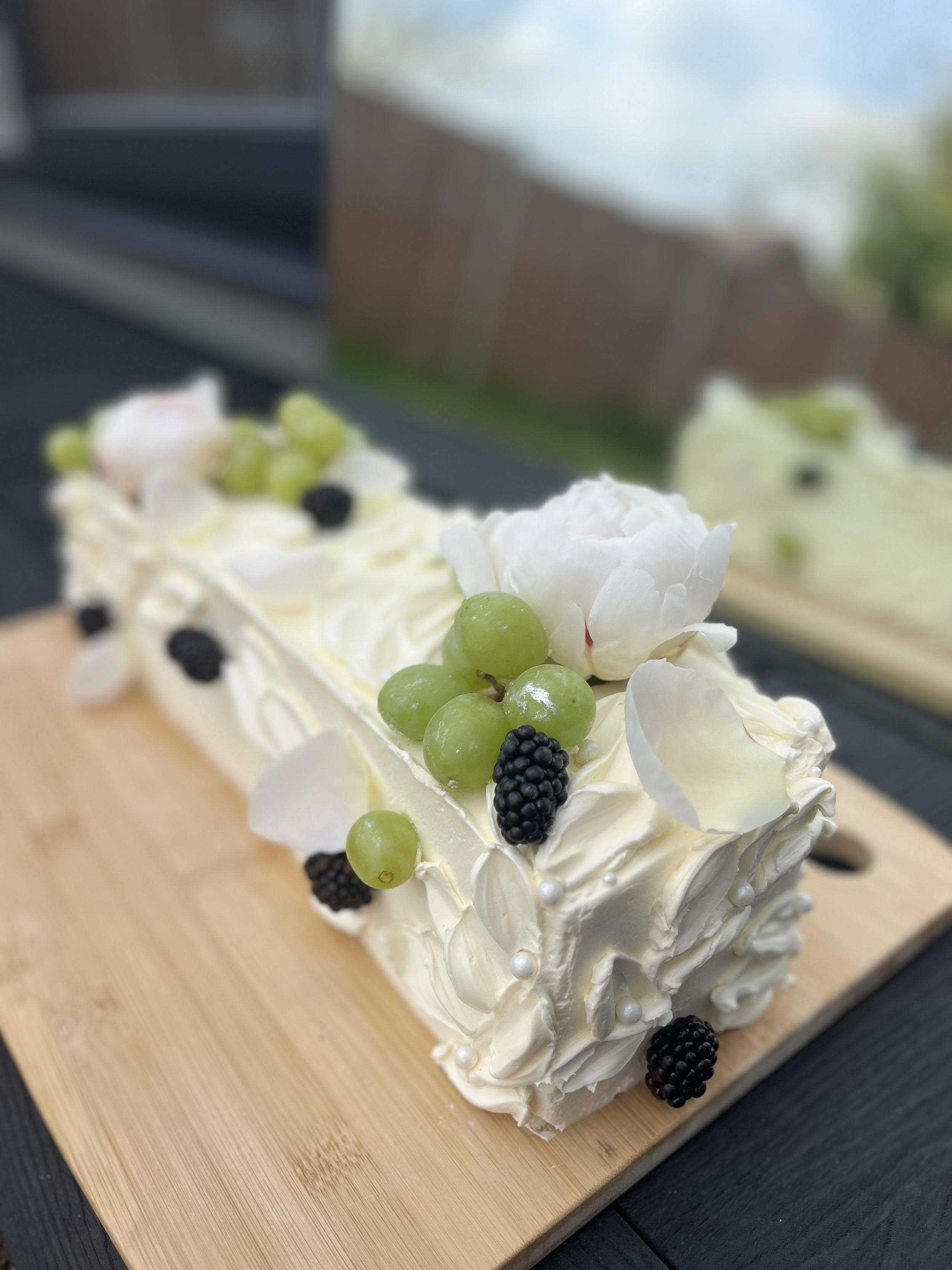 A rectangular white cake decorated with green grapes, blackberries, white flower petals, and pearl-like sugar decorations, placed on a wooden board outdoors.