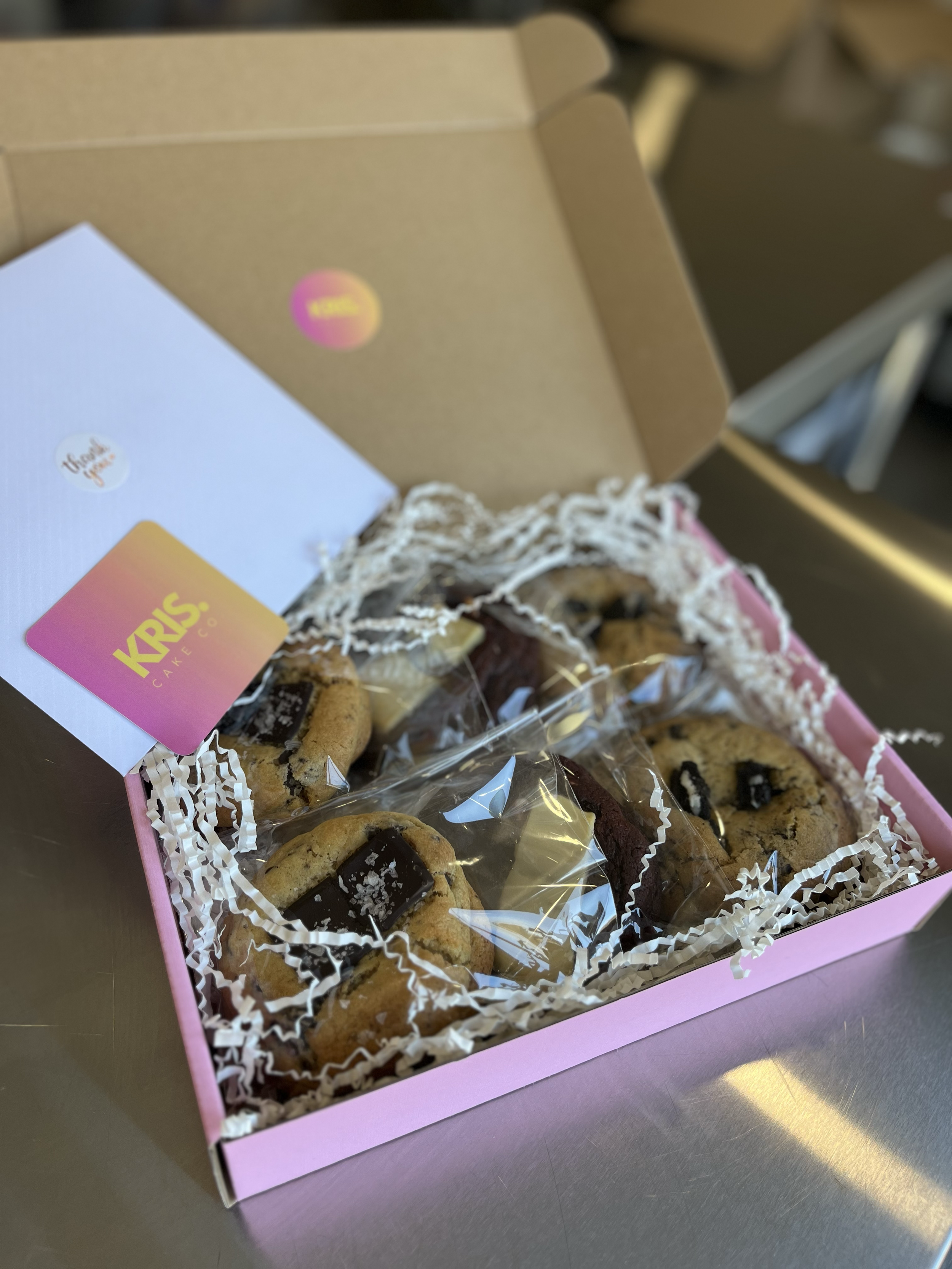 Pink box with assorted cookies wrapped in plastic, with white crinkle paper inside, on a metal surface.
