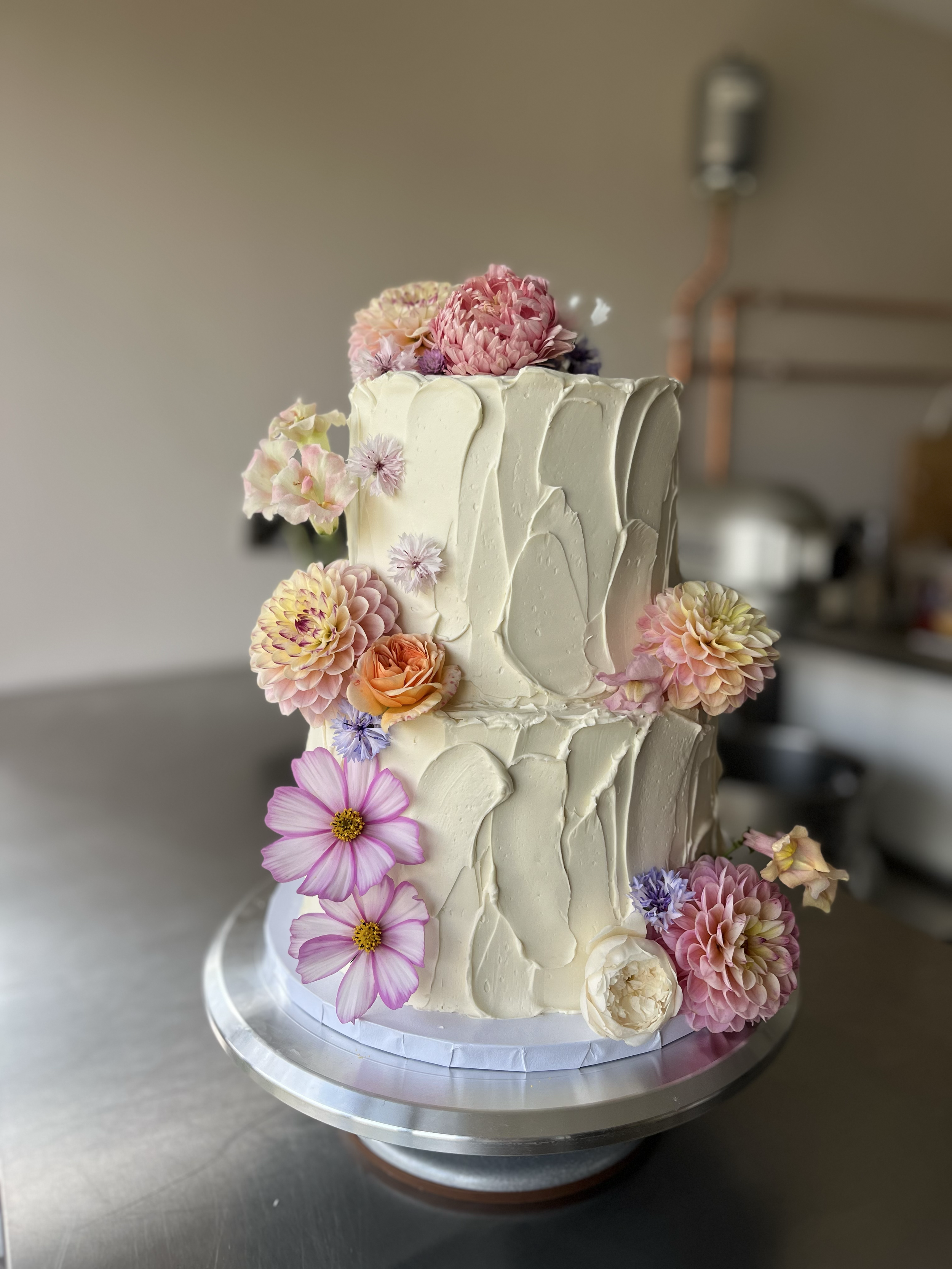 Two-tier white wedding cake decorated with assorted pink, purple, peach, and cream flowers, including dahlias, roses, and cosmos, on a silver cake stand.