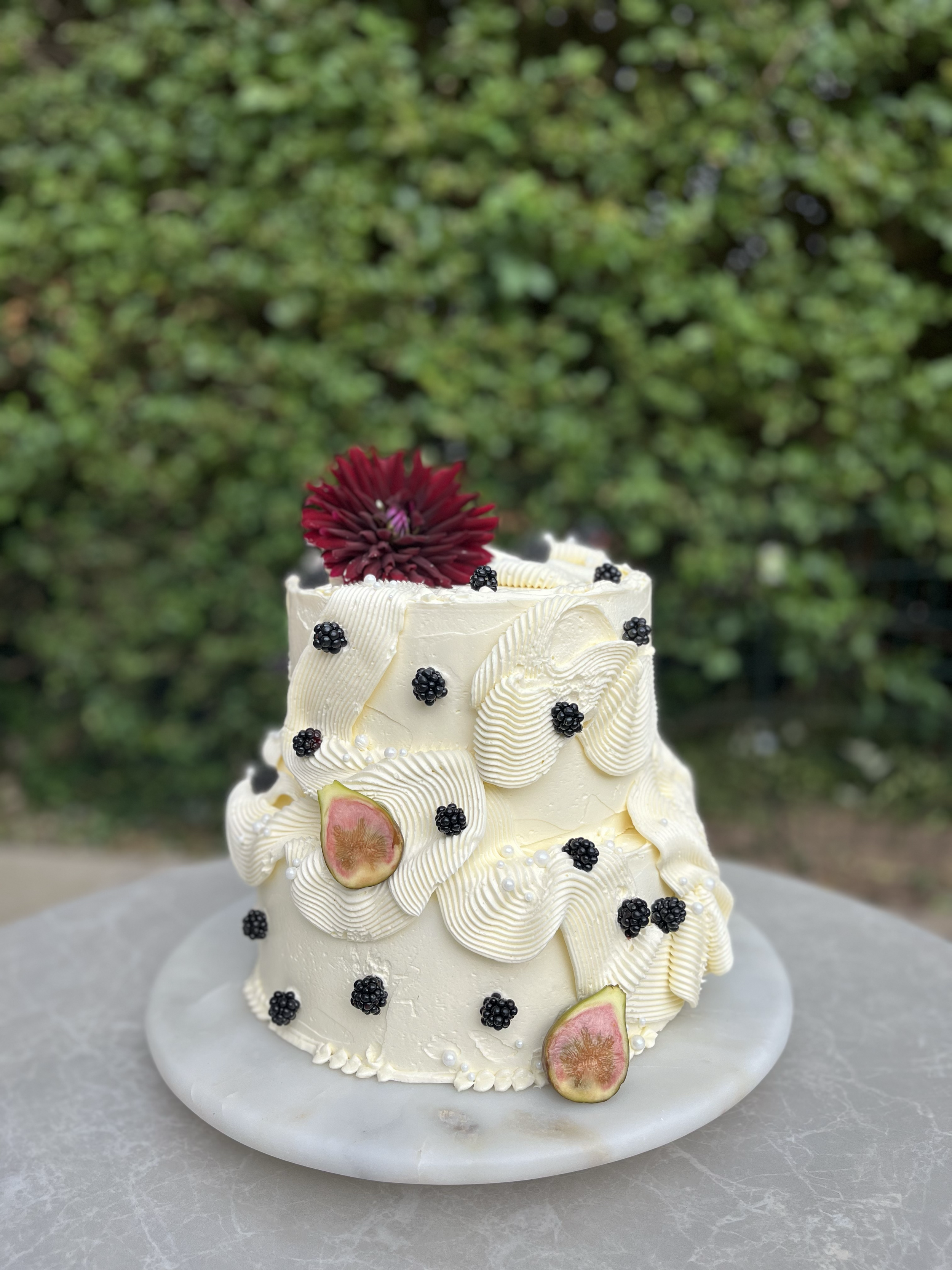A white frosted cake decorated with black berries, a sliced fig, white piping, and a large dark red flower on top, placed on a white marble cake stand outdoors.