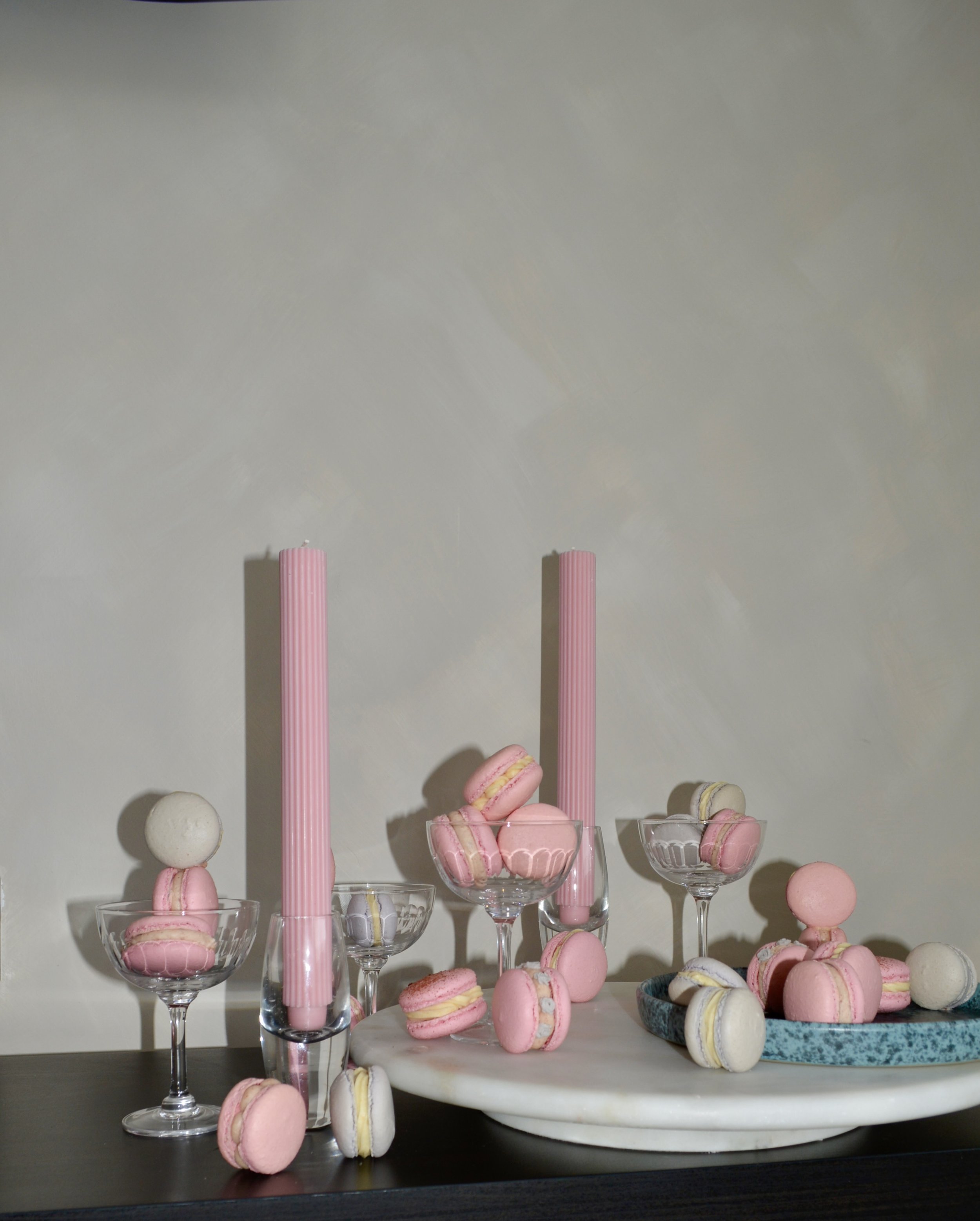Pink and white macarons displayed in glass cups and on a blue tray, with tall pink candles and a white marble serving board on a dark table against a plain wall.