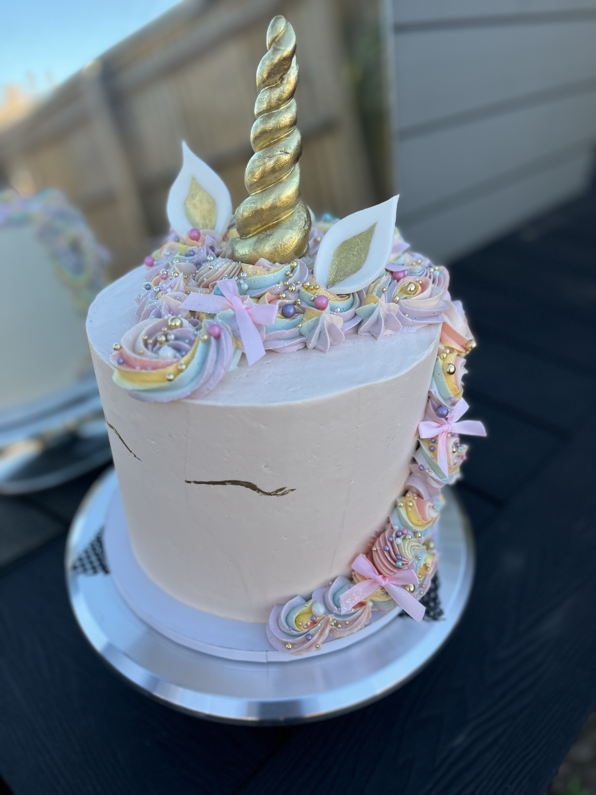 A pastel-colored unicorn-themed cake decorated with a golden spiral horn, white ears with gold accents, and colorful swirled icing with sprinkles and pink ribbons.