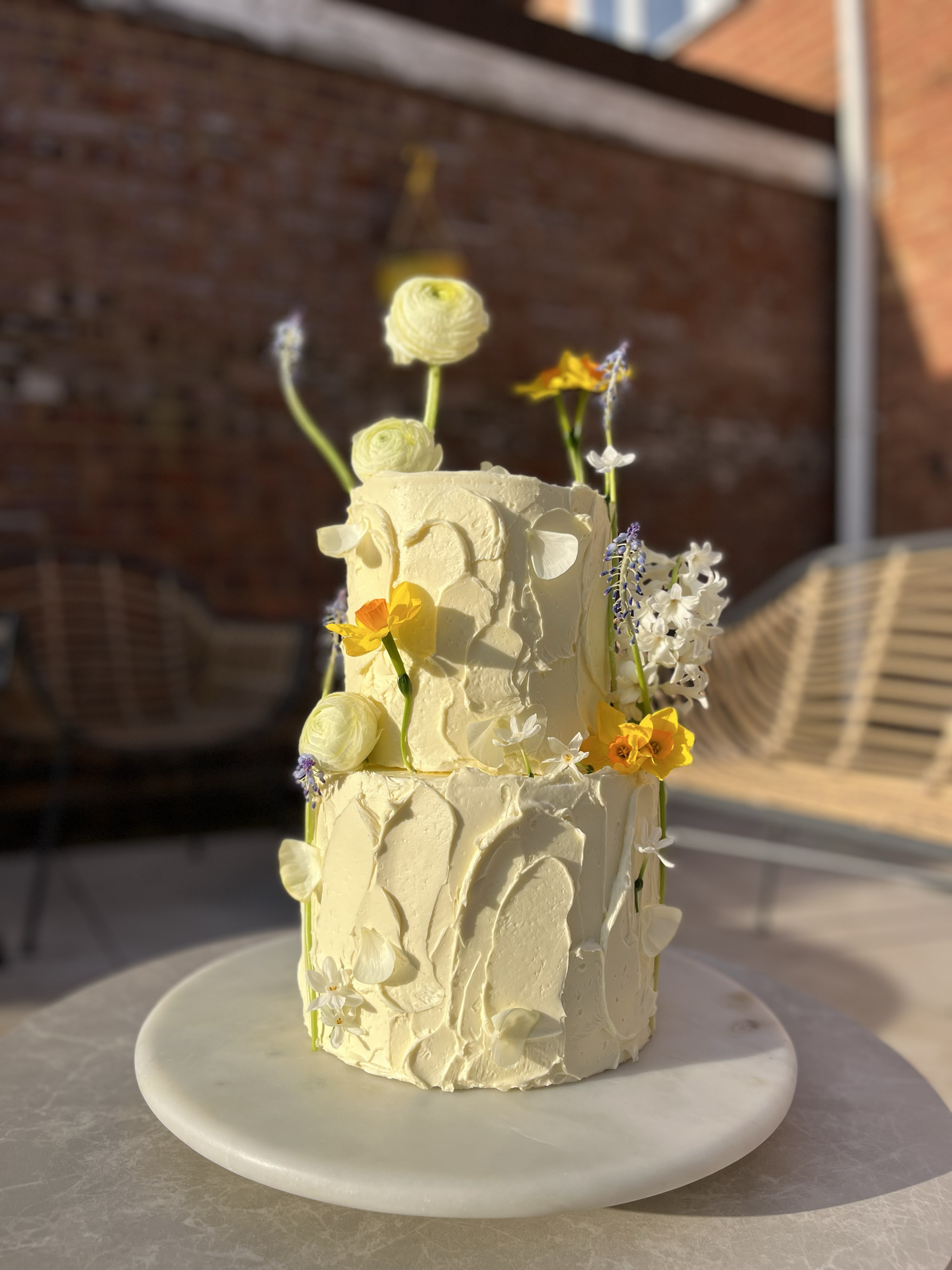 Two-tier white cake decorated with white and yellow flowers, placed on a white marble stand outside with a brick wall and wooden furniture in the background.