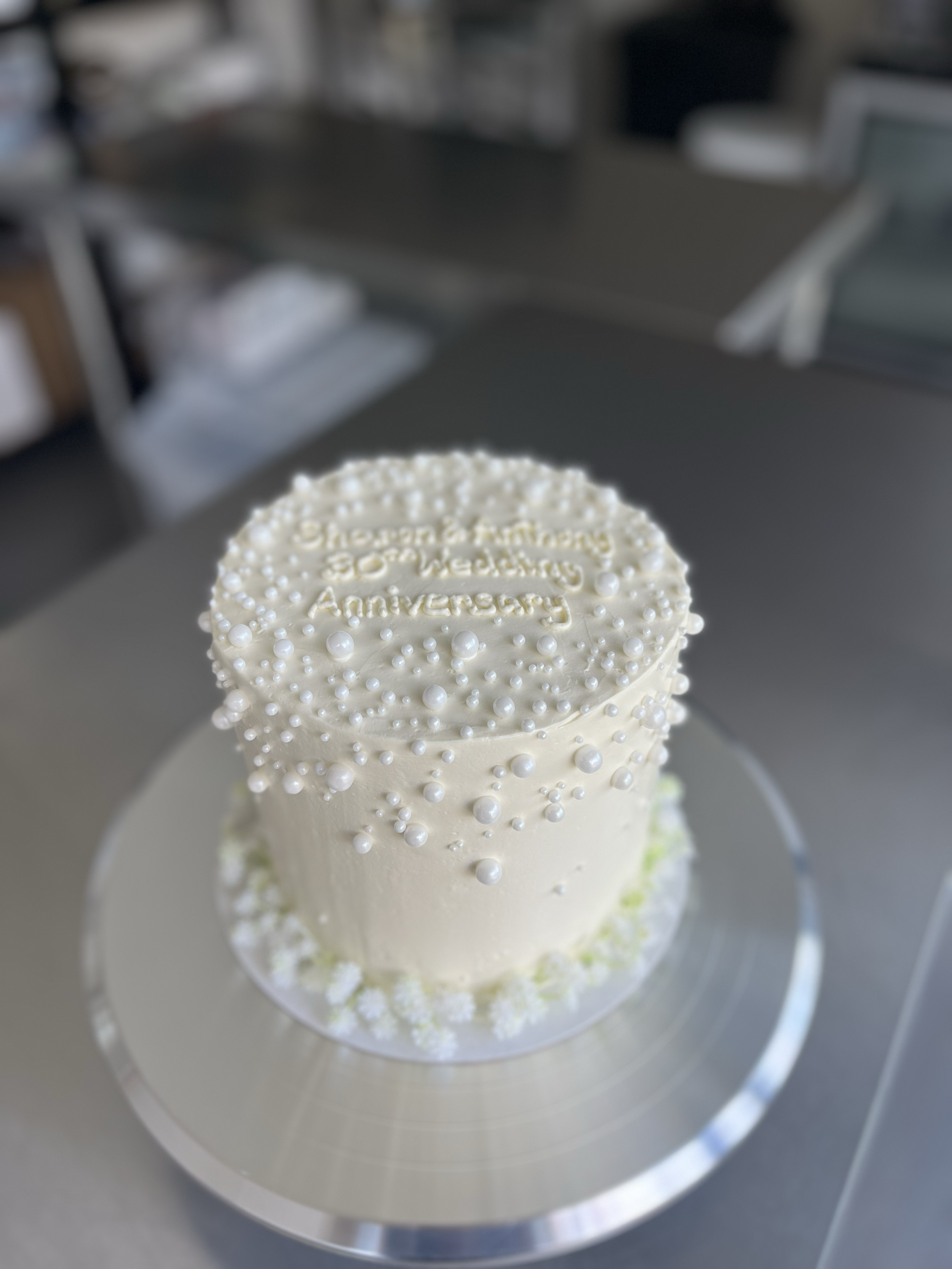 A white anniversary cake decorated with small white pearls and floral accents, with a message inscribed on top.